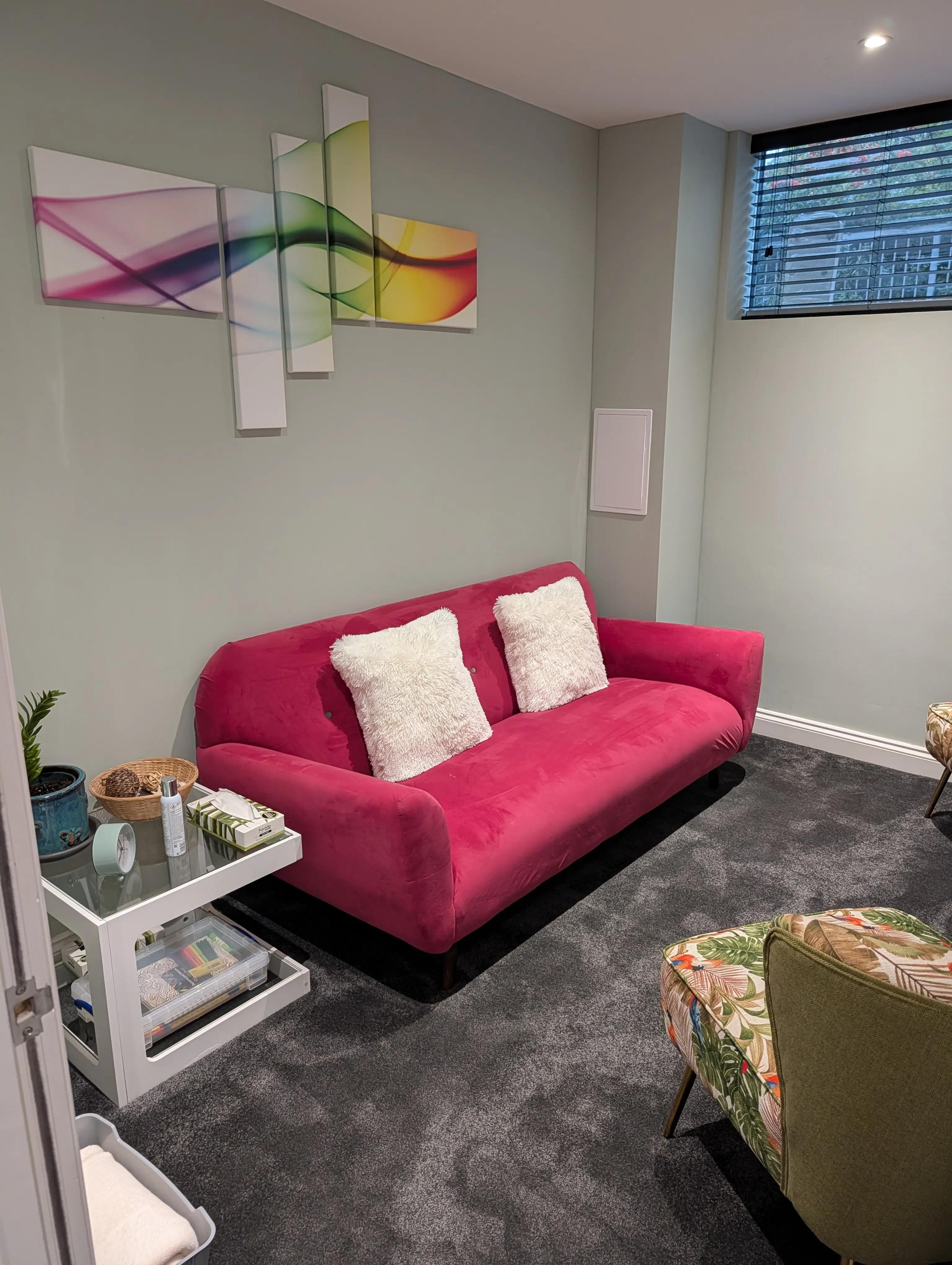A second Chelmsford therapy room, with a pink sofa and cushions, an armchair, a side table with tissues and abstract canvases on a sage wall