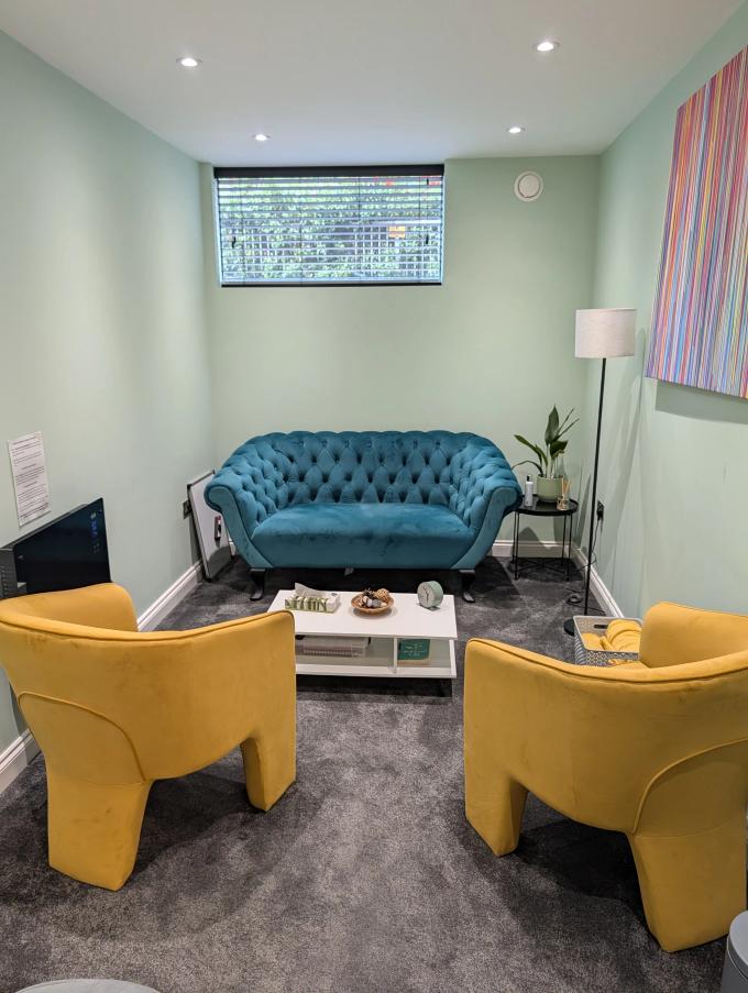 A Chelmsford therapy room set up for a session: two mustard armchairs facing a teal sofa across a low table, with a window and a floor lamp