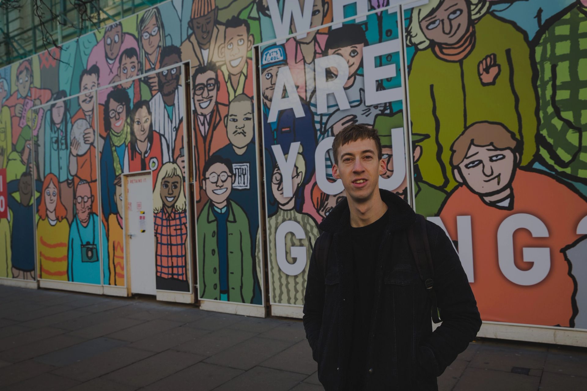 A smiling young man stands on a street in front of a colorful mural depicting diverse cartoon figures and the text "WHAT ARE YOU GOING".