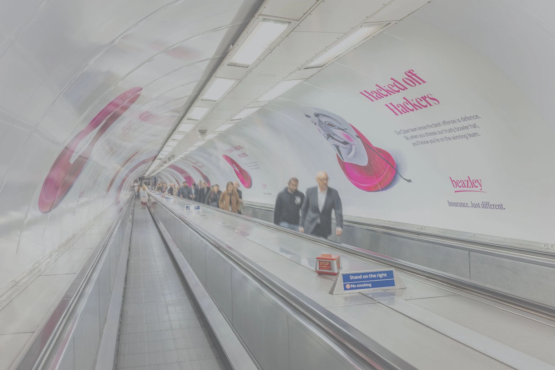 A long, curving subway escalator tunnel with people riding up, featuring a "Hacked off Hackers" ad for Beazley Insurance on the wall.