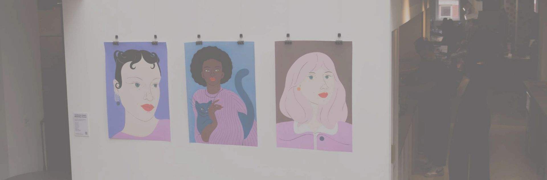 Three colorful portraits of women, one holding a cat, hanging on a white wall.
