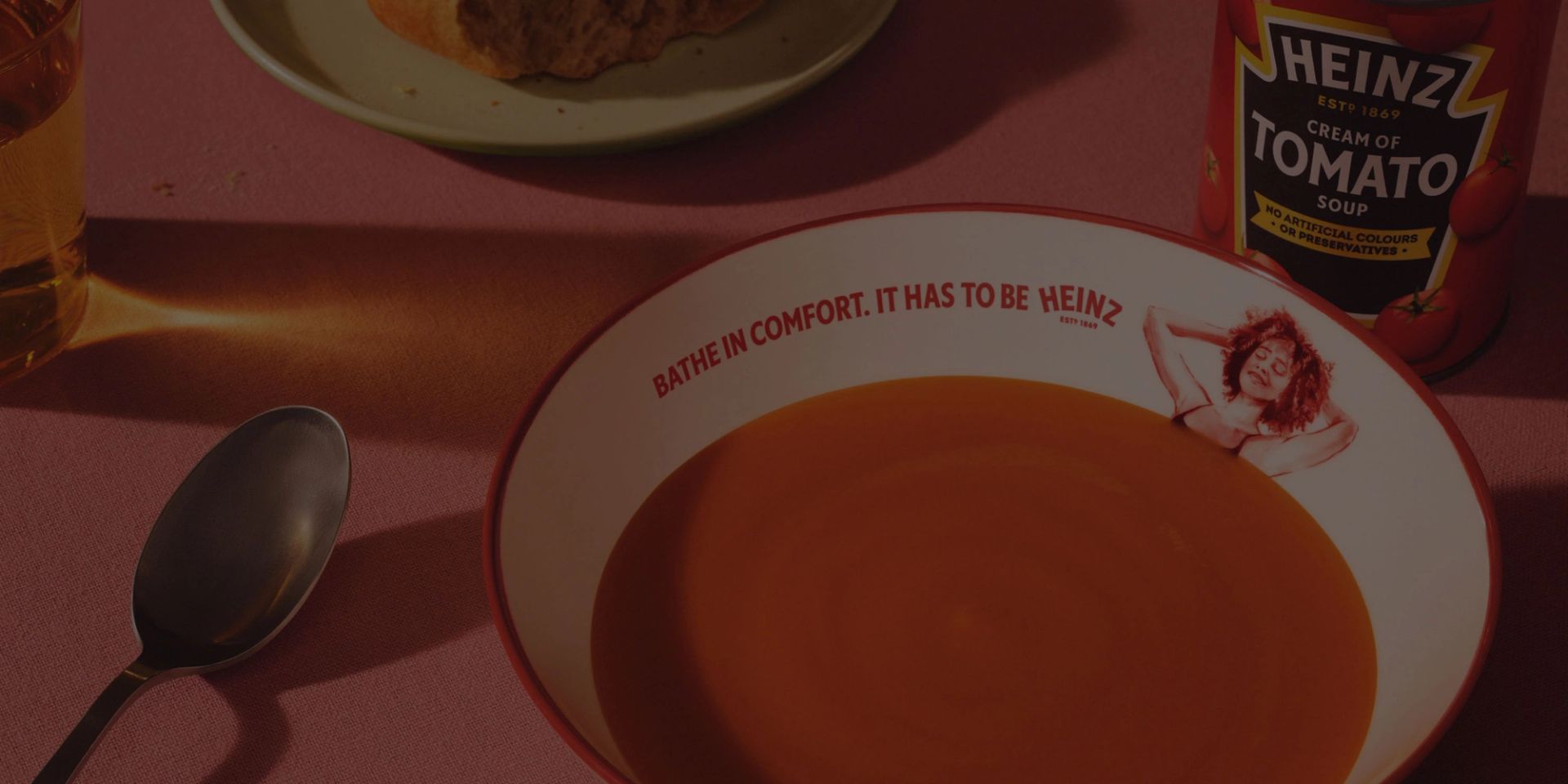 A bowl of tomato soup, a spoon, a glass, and a Heinz soup can on a pink table. The bowl has "BATHE IN COMFORT. IT HAS TO BE HEINZ" and an illustration of a woman relaxing.