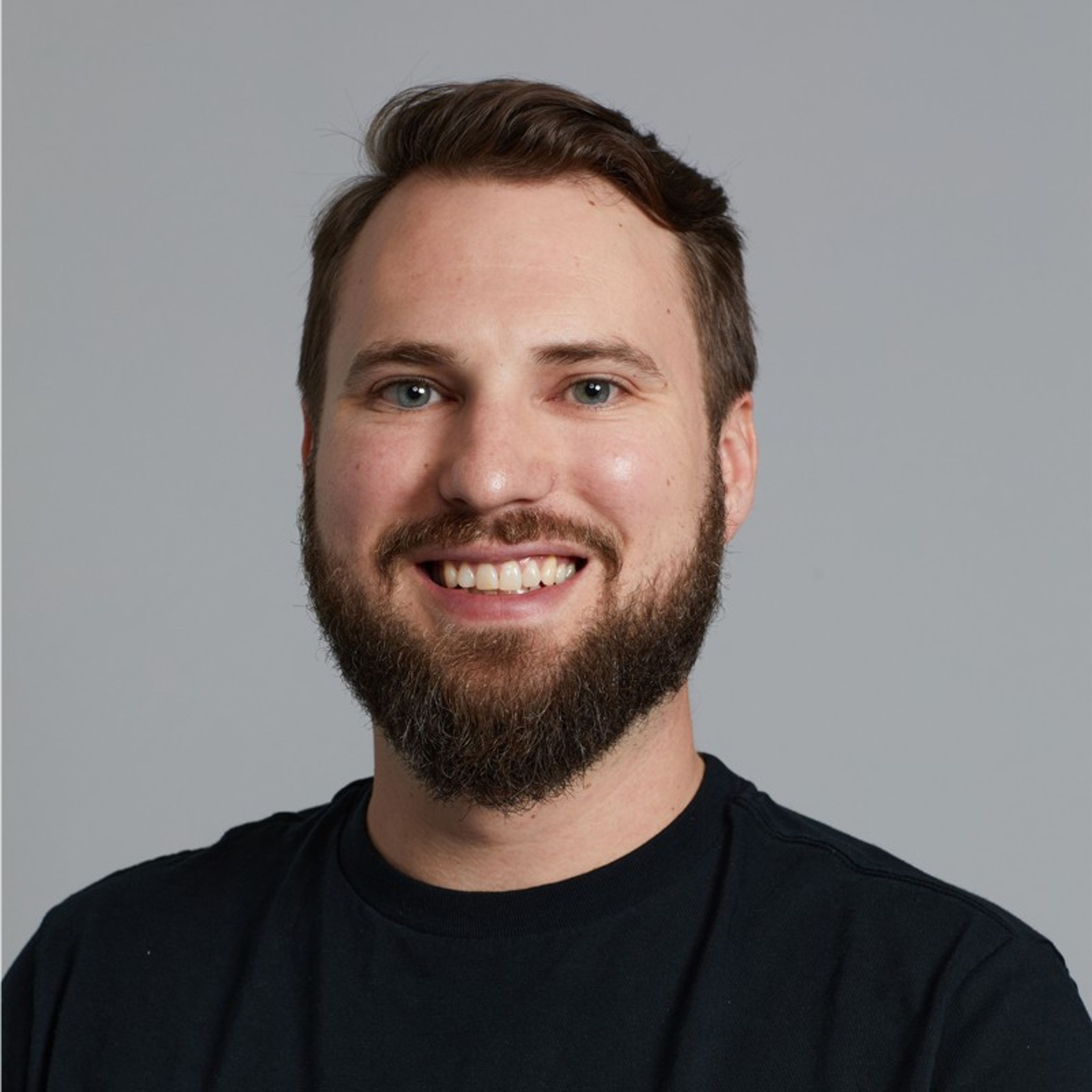 Headshot of a smiling man with a beard and short dark hair against a gray background.