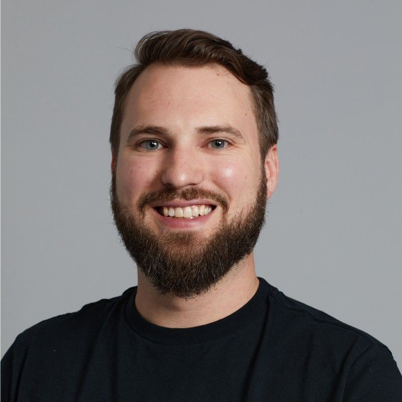 Headshot of a smiling man with a beard and short dark hair against a gray background.