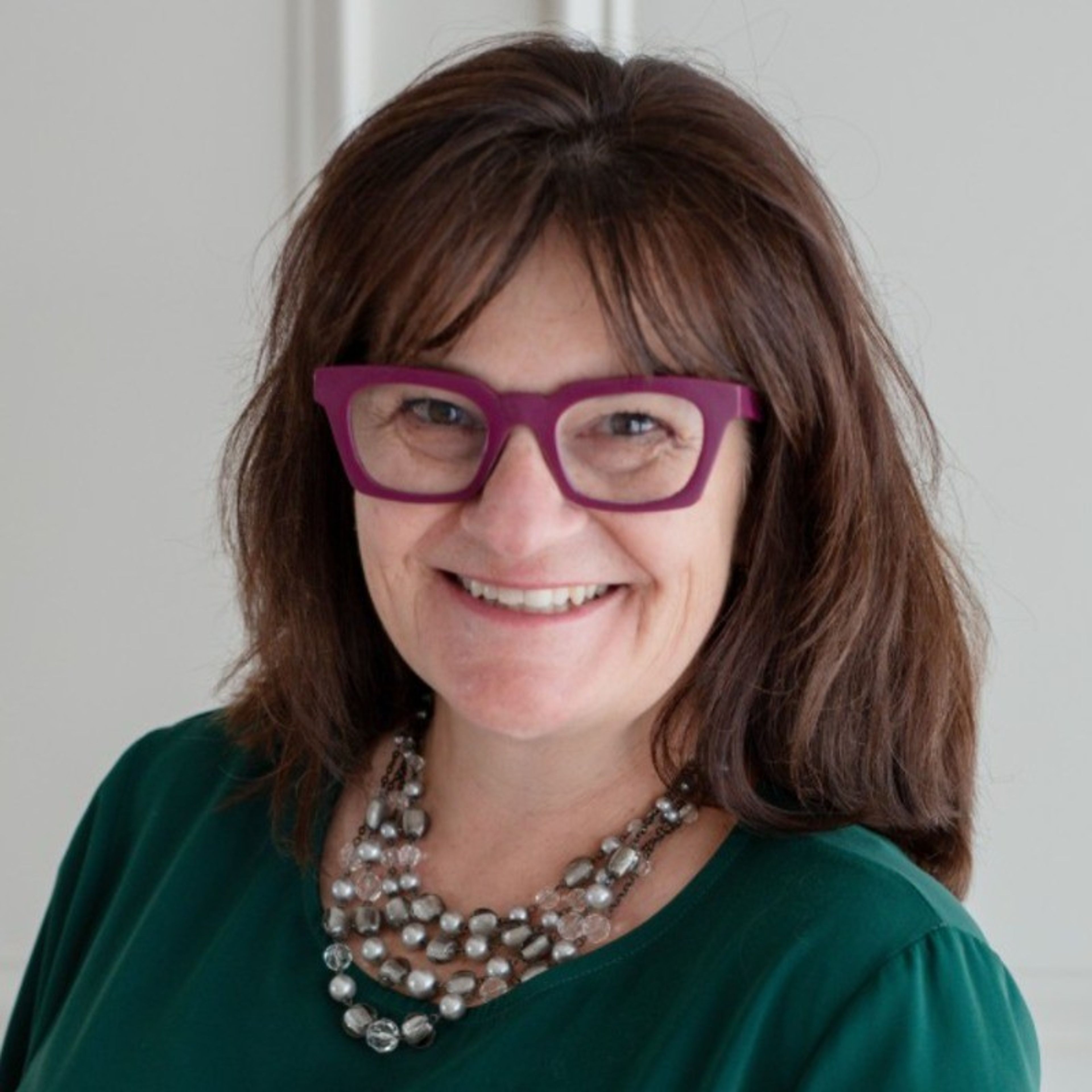 A smiling woman with brown hair, vibrant purple glasses, and a multi-strand beaded necklace.