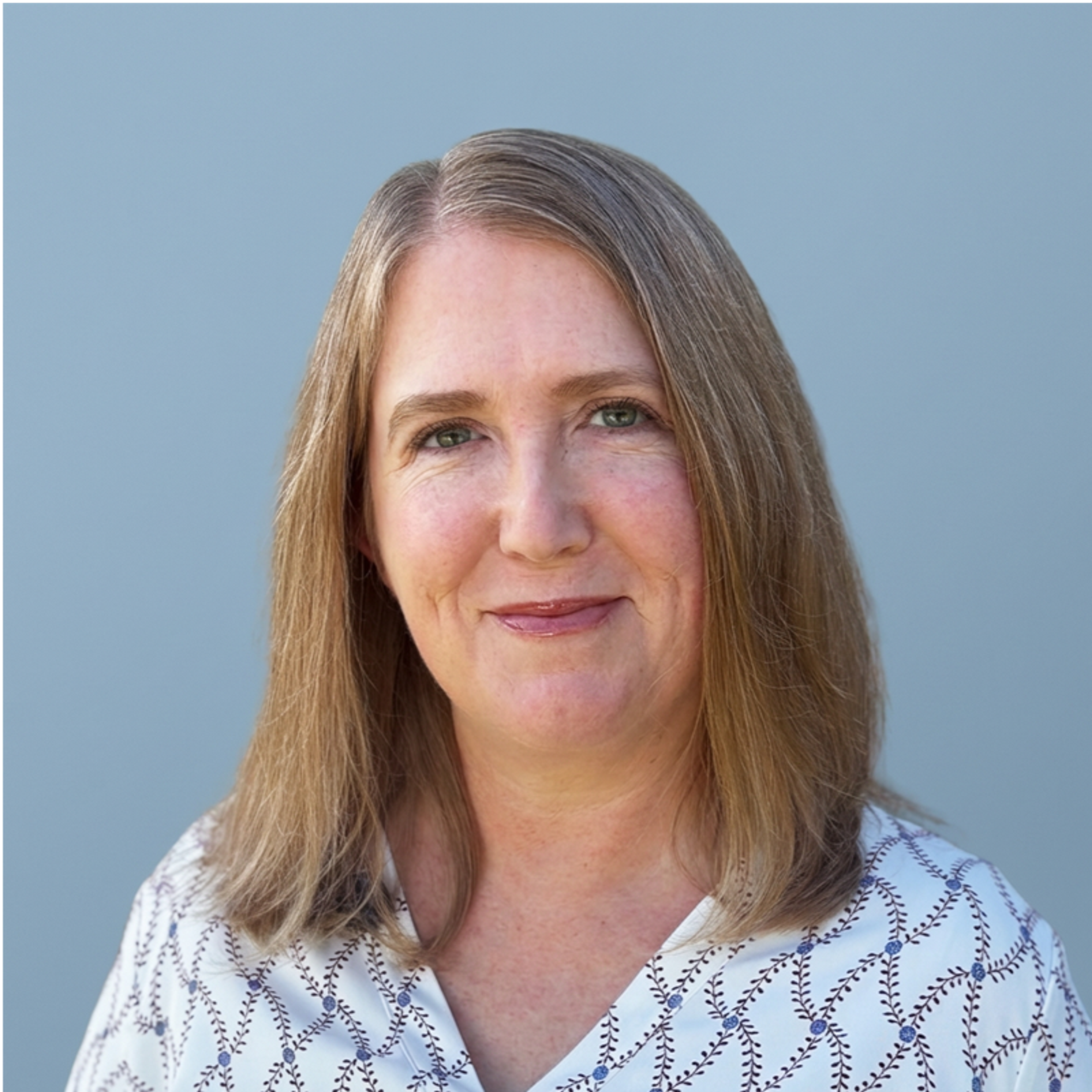 A smiling woman with shoulder-length blonde hair and green eyes wears a patterned shirt against a light blue background.
