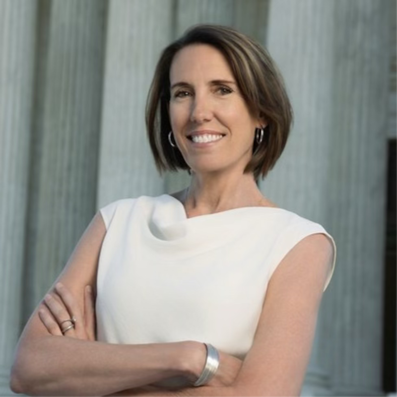 Smiling woman with short brown hair and white top, arms crossed, in front of columns.