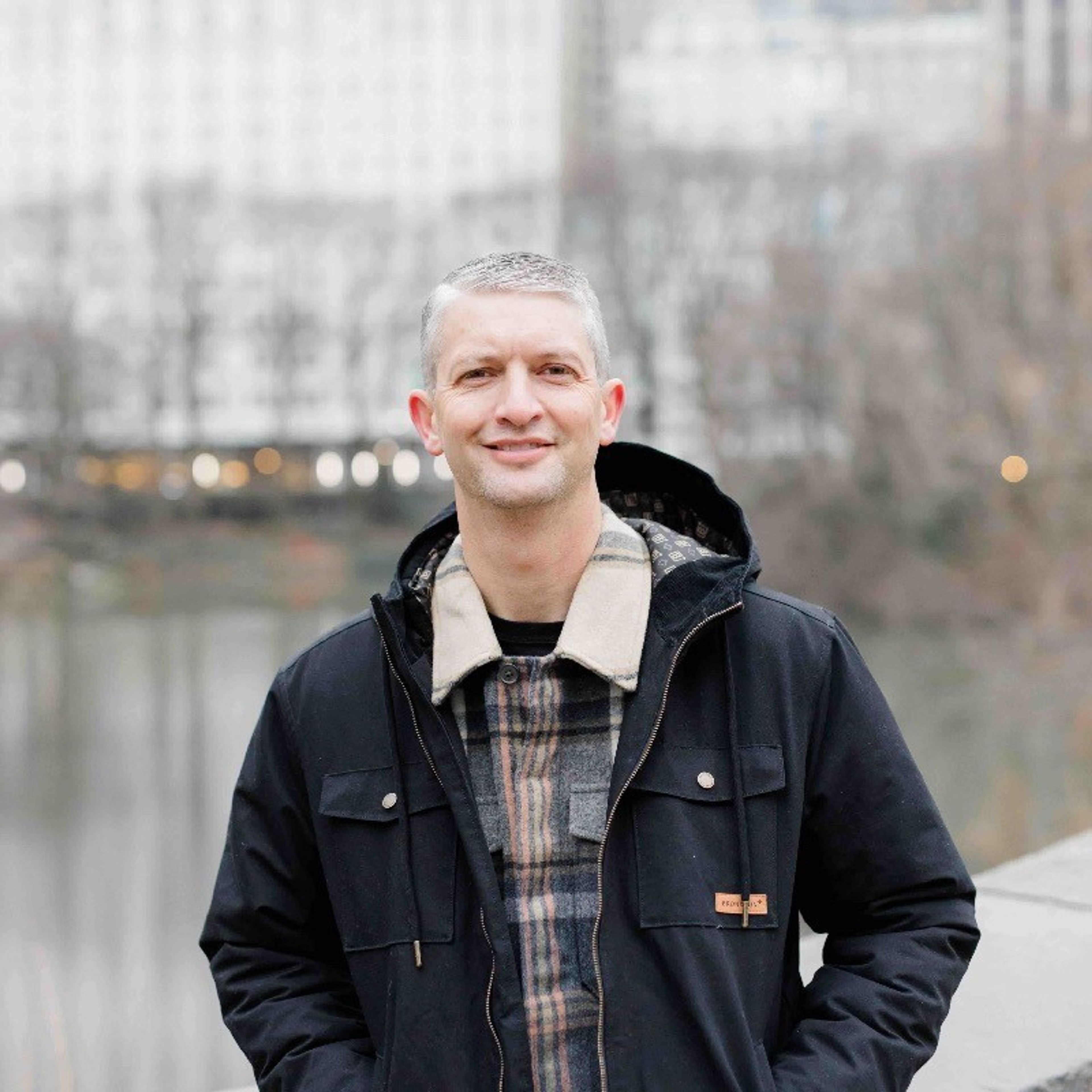 Smiling man with gray hair wearing a dark jacket and plaid shirt outdoors.