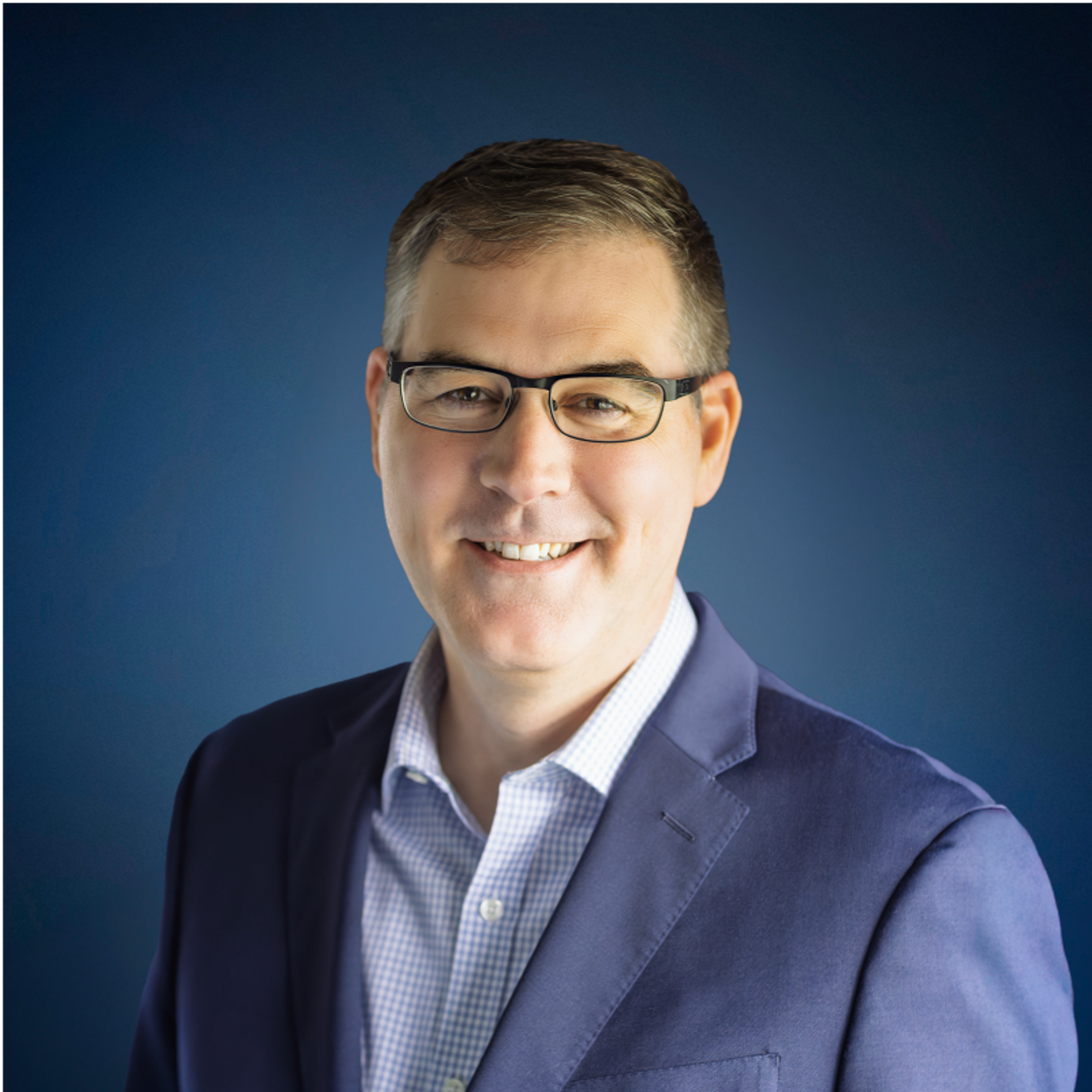 A smiling man with glasses in a blue suit jacket and light blue shirt against a blue background.
