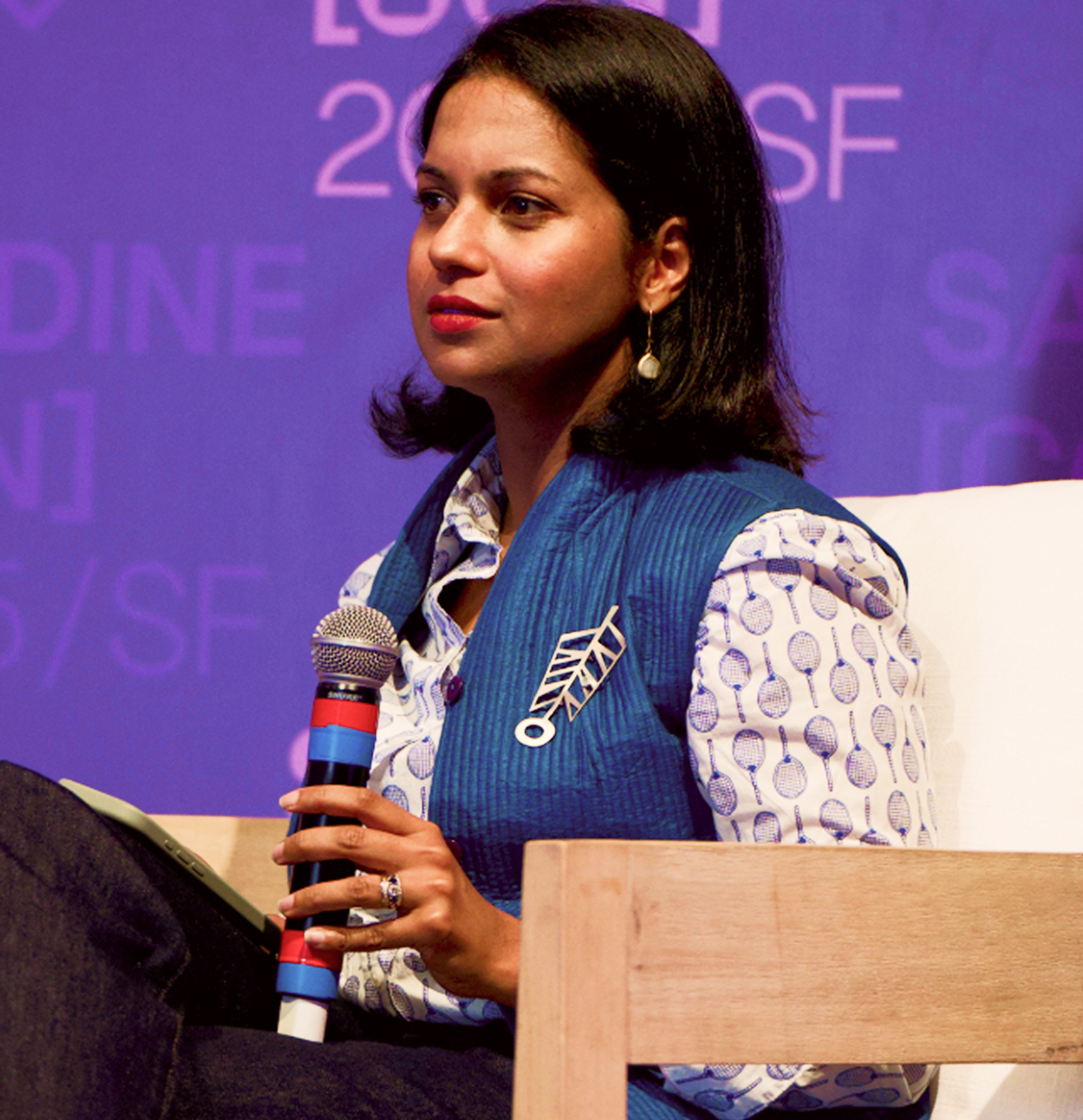 A woman with dark hair and red lipstick wears a blue vest over a patterned shirt with a brooch, holding a microphone and looking to her right.
