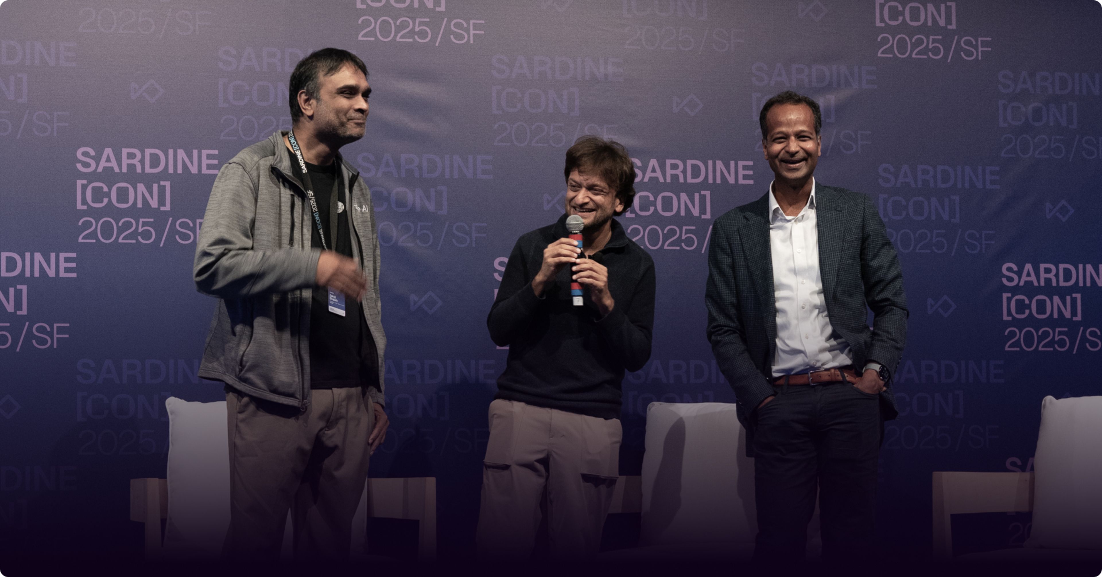 Three smiling men on stage at SardineCon, one holding a microphone.