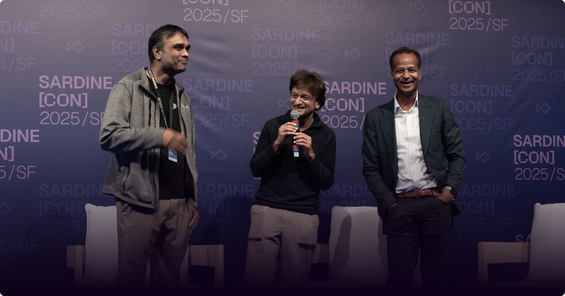Three smiling men on stage at SardineCon, one holding a microphone.