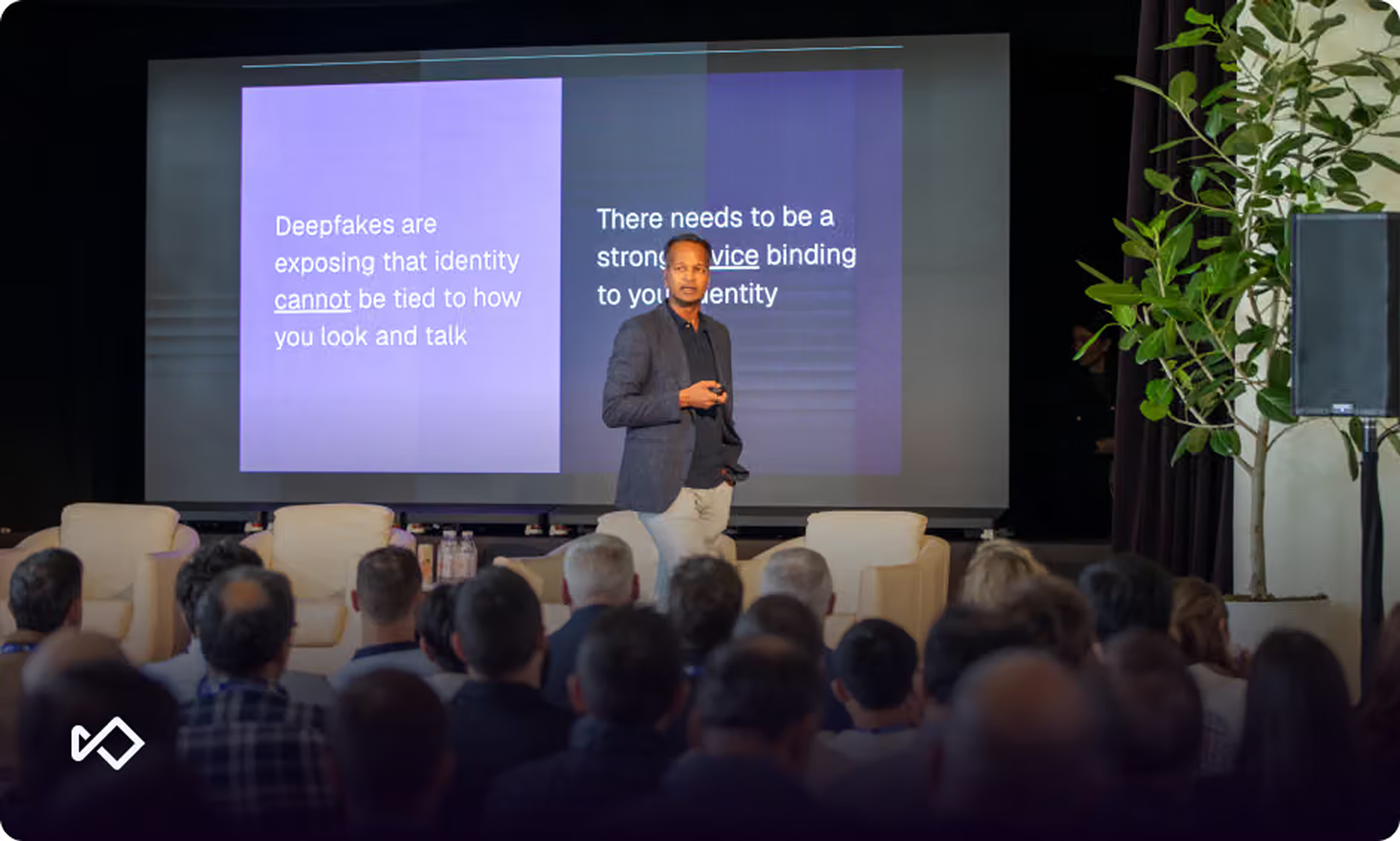 A man presents on stage about deepfakes and identity on a large screen to a seated audience.
