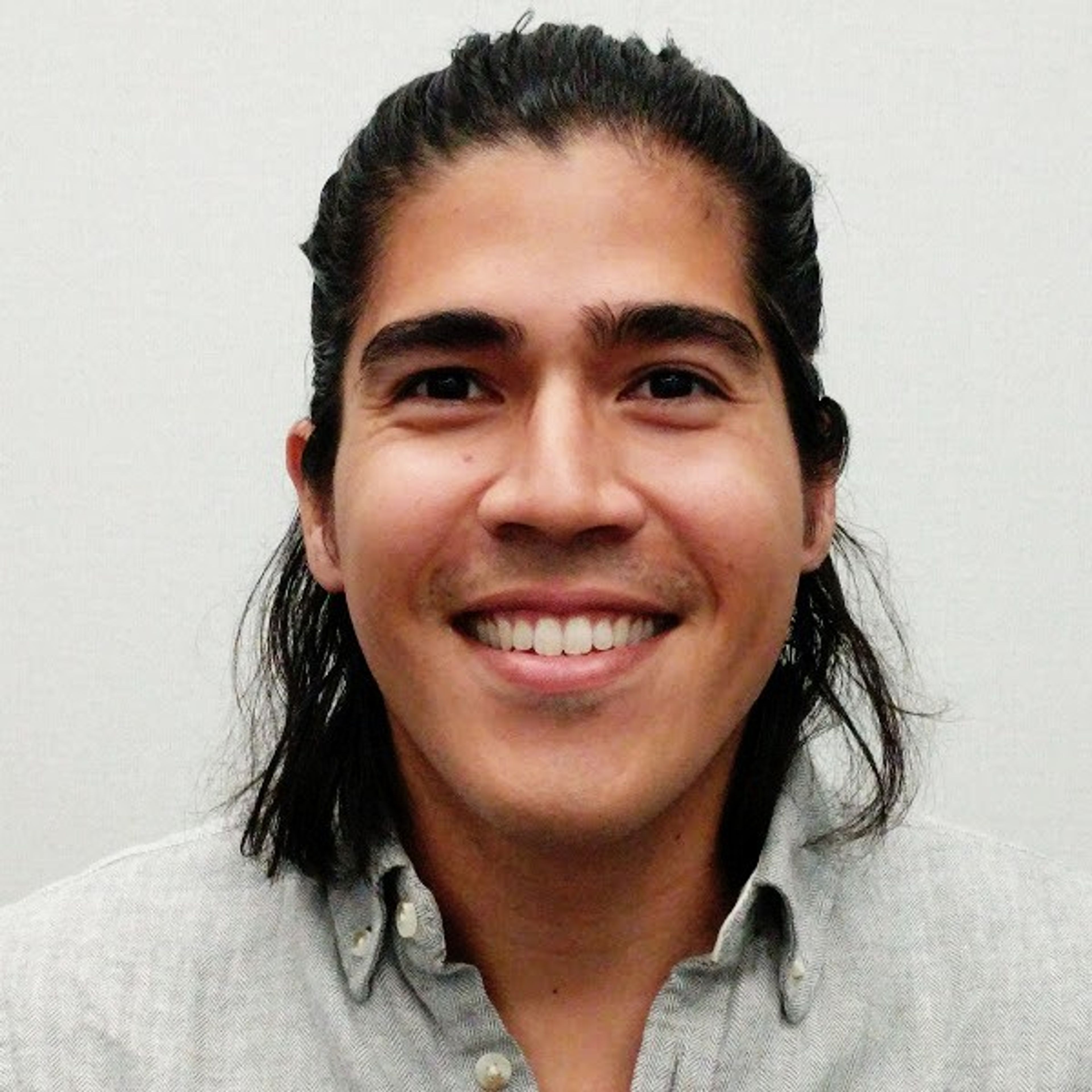 A man with long dark hair pulled back, smiling and wearing a grey collared shirt.