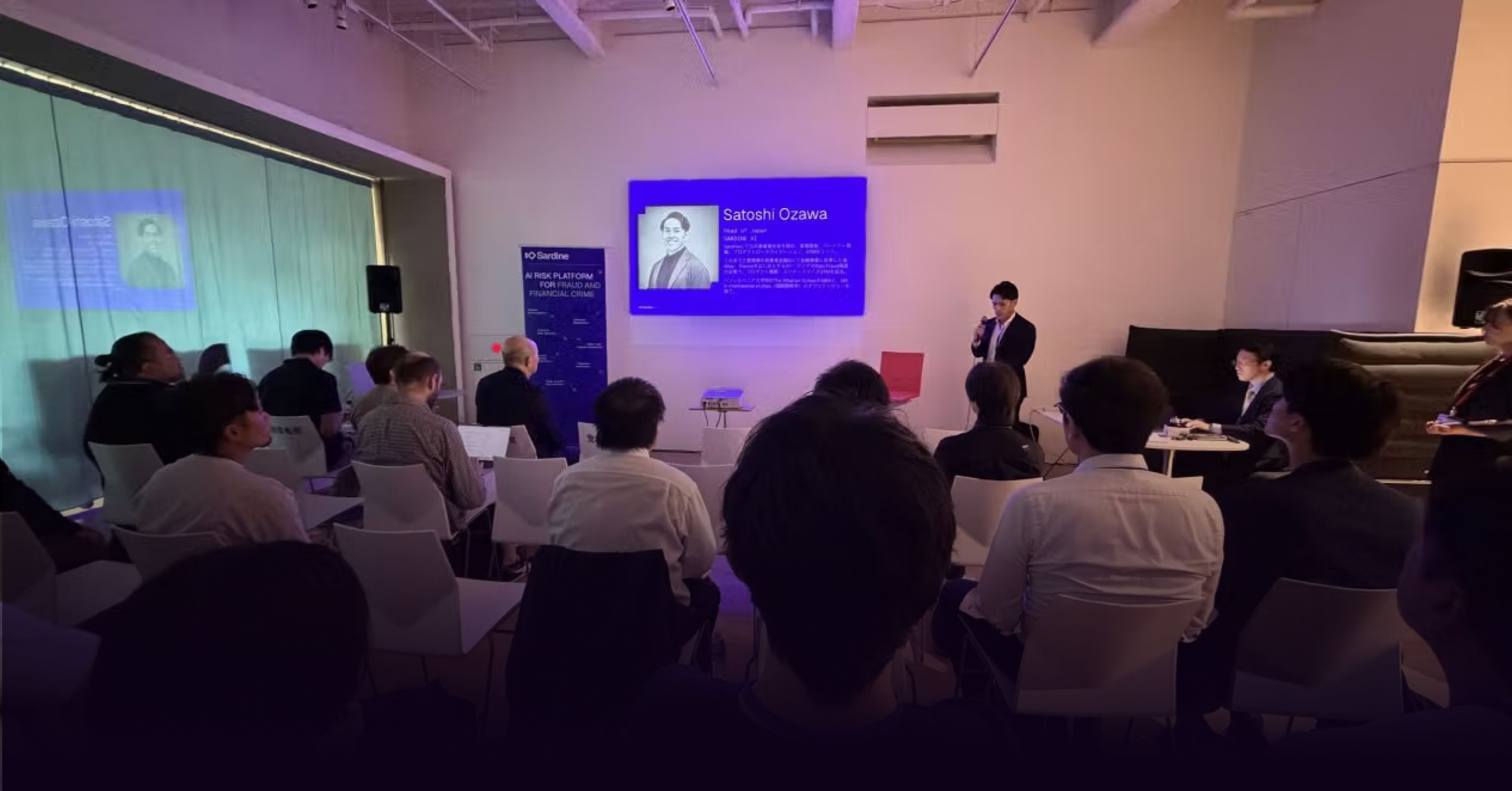 A speaker addresses an audience in a room with screens displaying "Satoshi Ozawa."