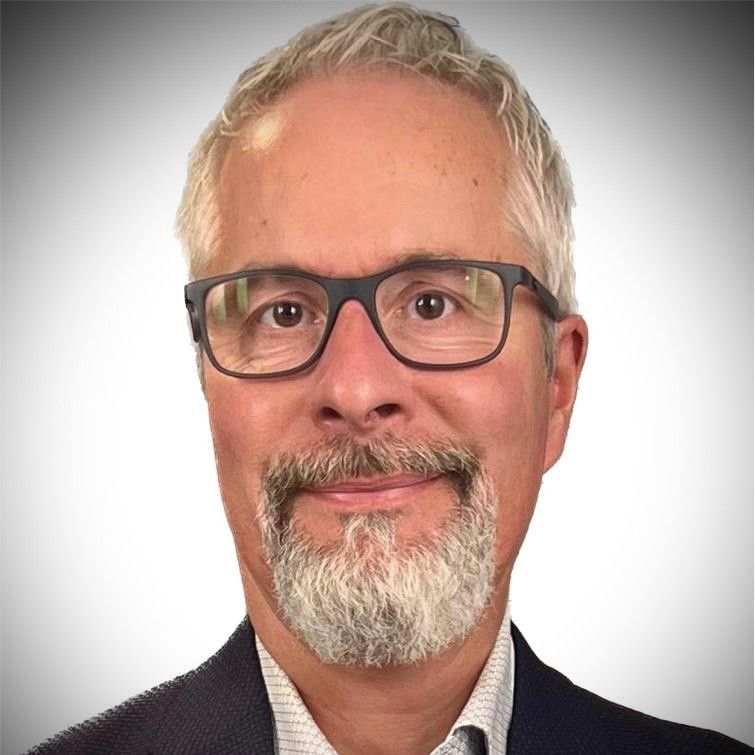 Headshot of a smiling man with gray hair, a gray beard, and black-rimmed glasses, wearing a dark blazer.