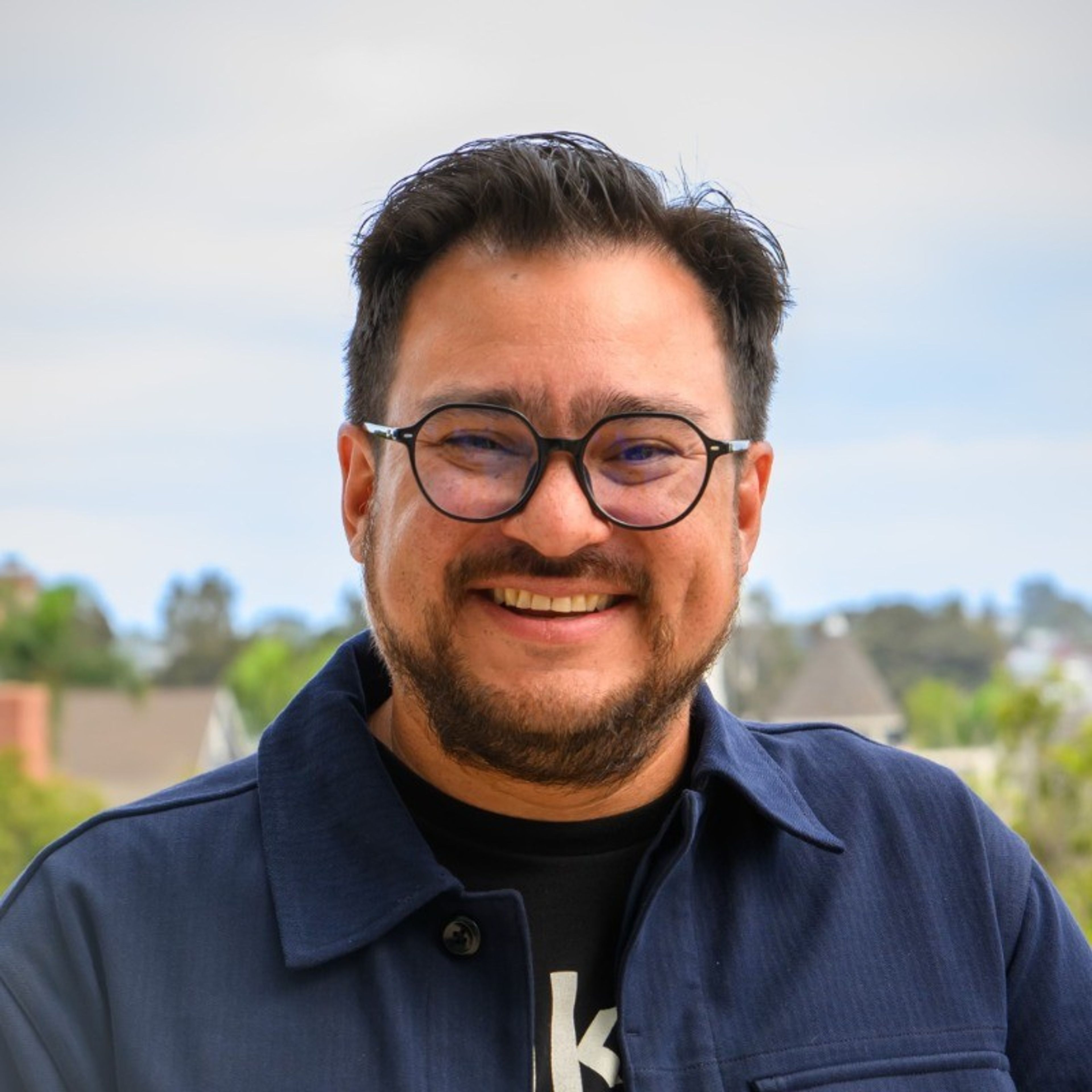A smiling man with glasses and a beard wears a dark blue jacket outdoors.