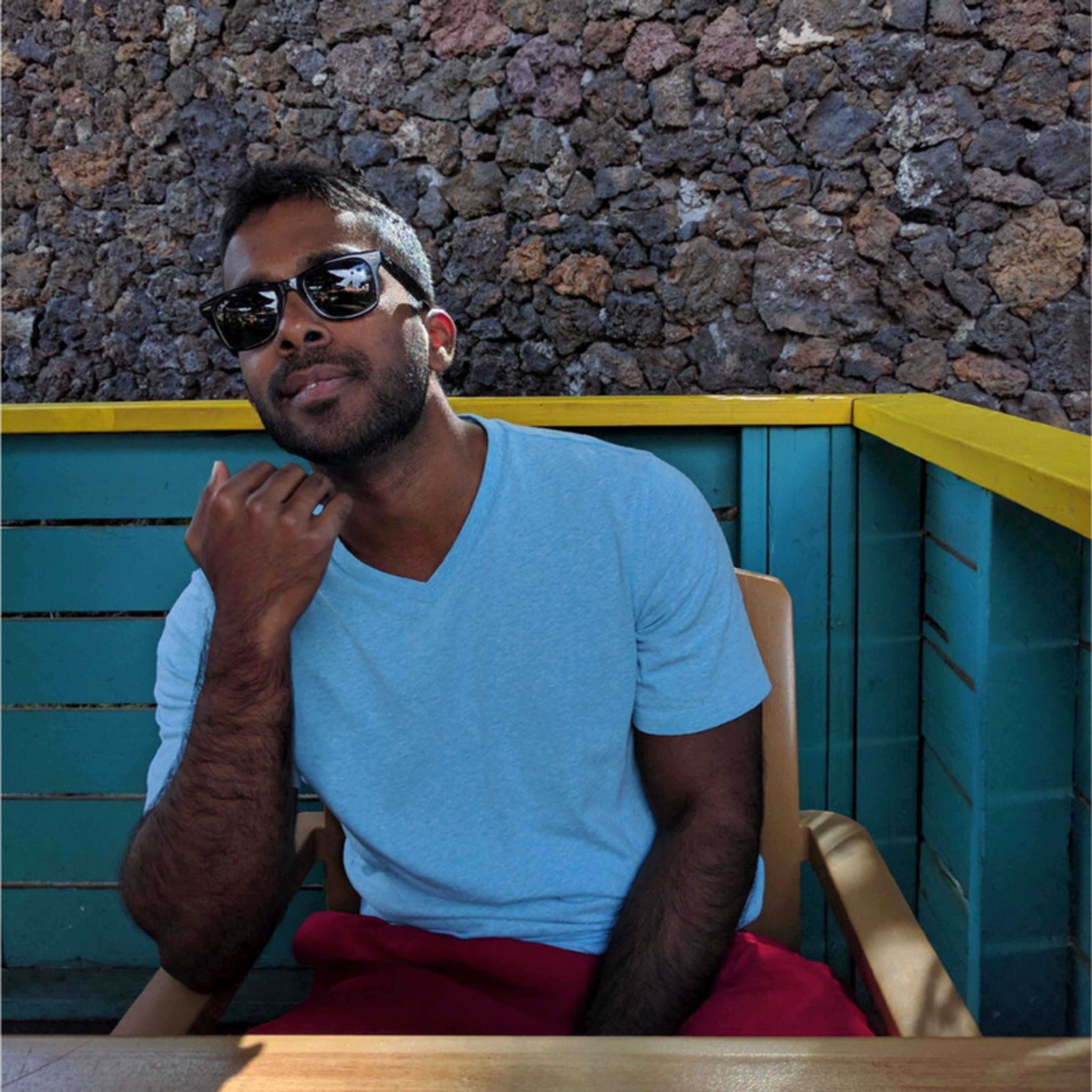 A man in sunglasses and a blue shirt sits in a chair, with a stone wall and blue railing behind him.