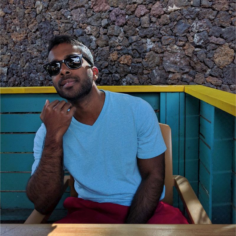 A man in sunglasses and a blue shirt sits in a chair, with a stone wall and blue railing behind him.