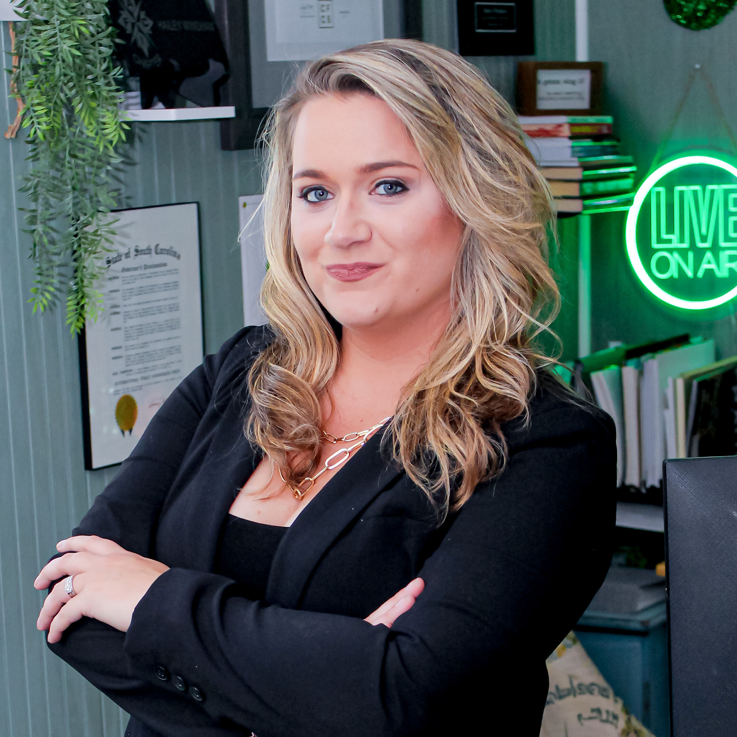 Smiling blonde woman in a black blazer with arms crossed, standing in a room with a "LIVE ON AIR" neon sign.
