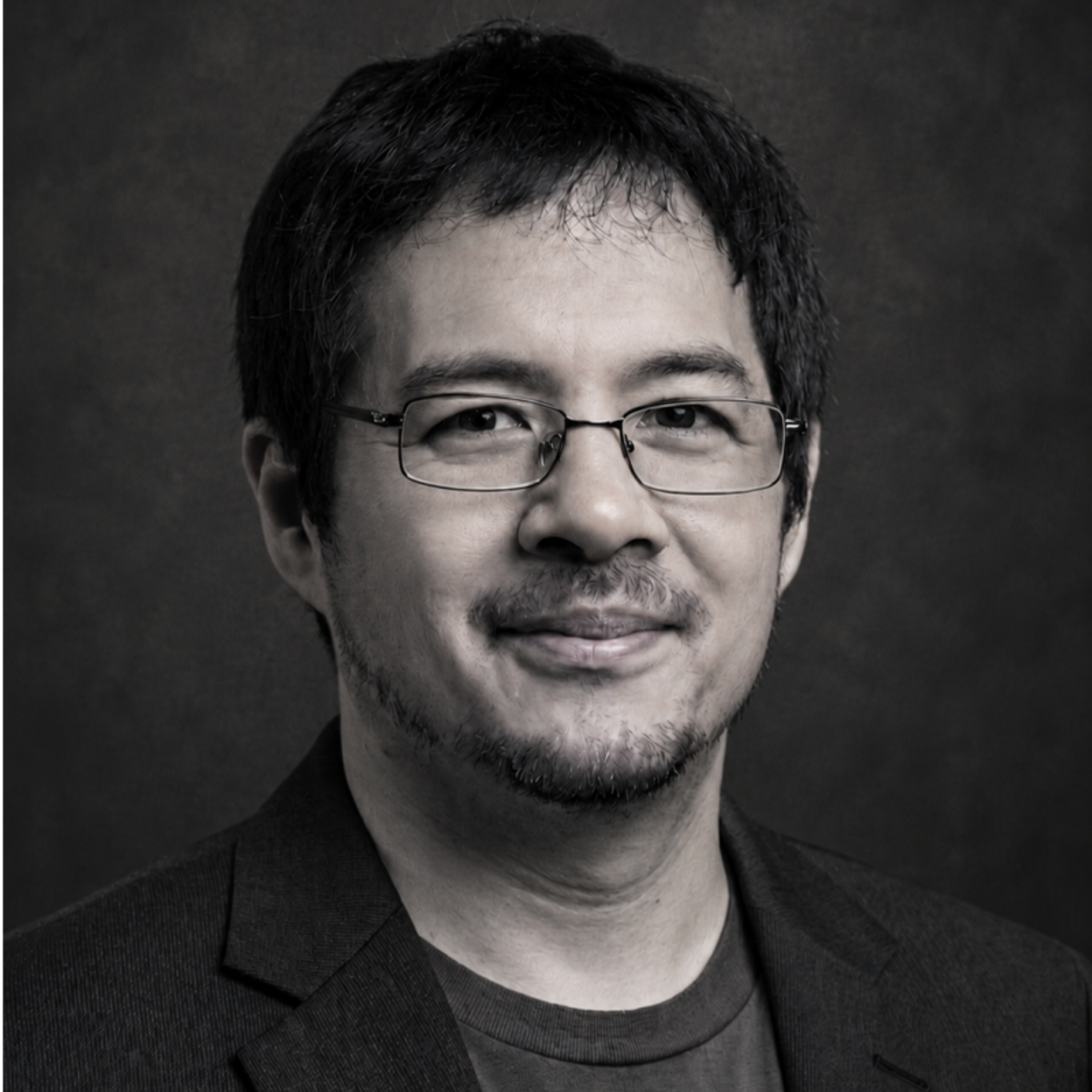 Black and white headshot of an Asian man with glasses, short dark hair, and a goatee, wearing a dark jacket and smiling.