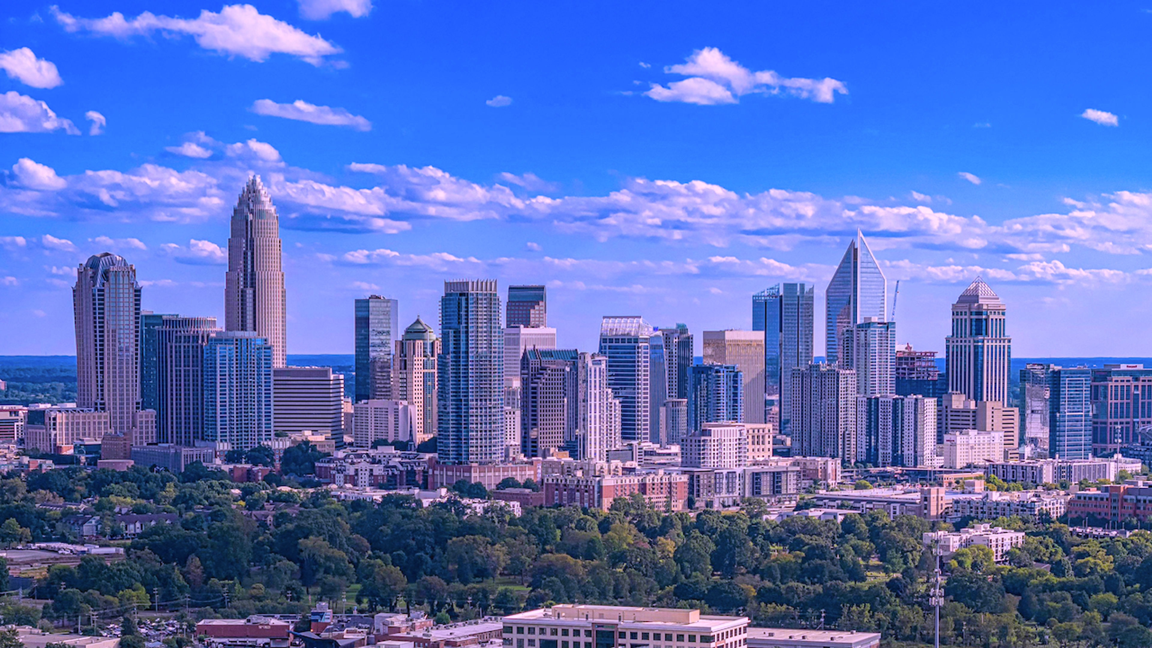 Charlotte, NC skyline with numerous skyscrapers, green trees, and a blue sky with clouds.