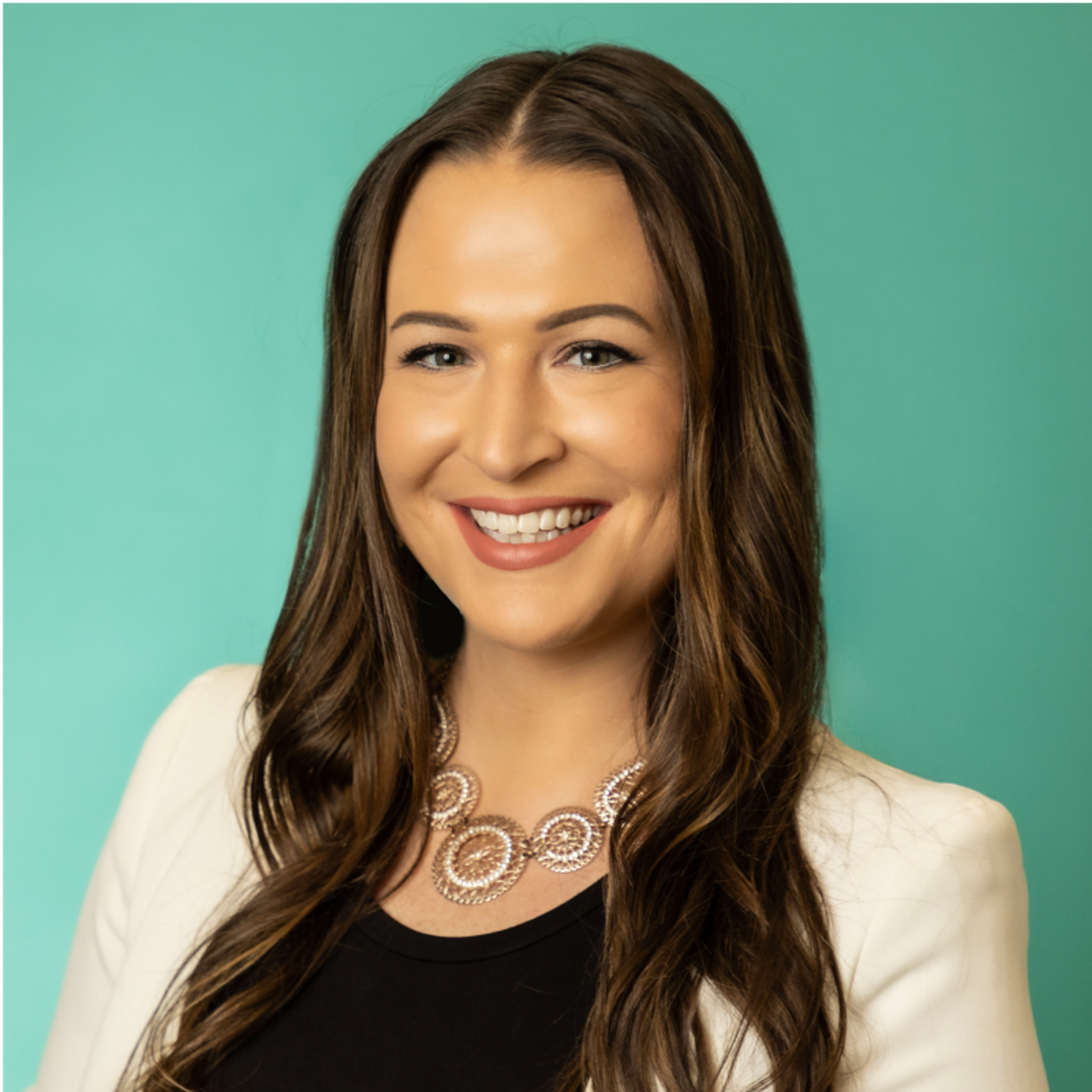 Headshot of a smiling woman with long brown hair, wearing a white blazer and a statement necklace against a teal background.