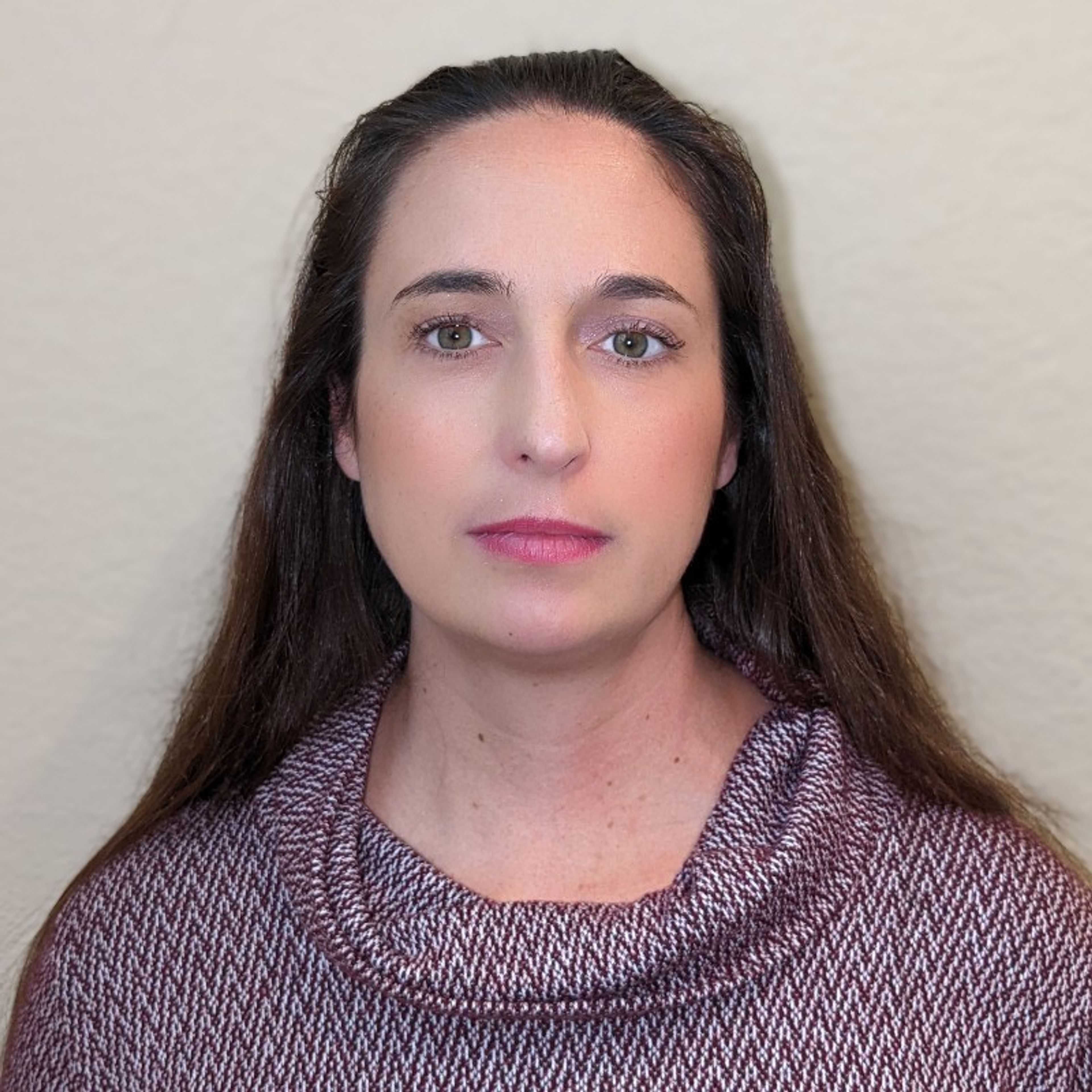A woman with long brown hair and green eyes, wearing a burgundy and white patterned cowl-neck sweater, looking directly forward.