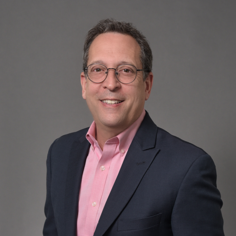A smiling man with graying hair and glasses, wearing a pink shirt and a dark blazer.