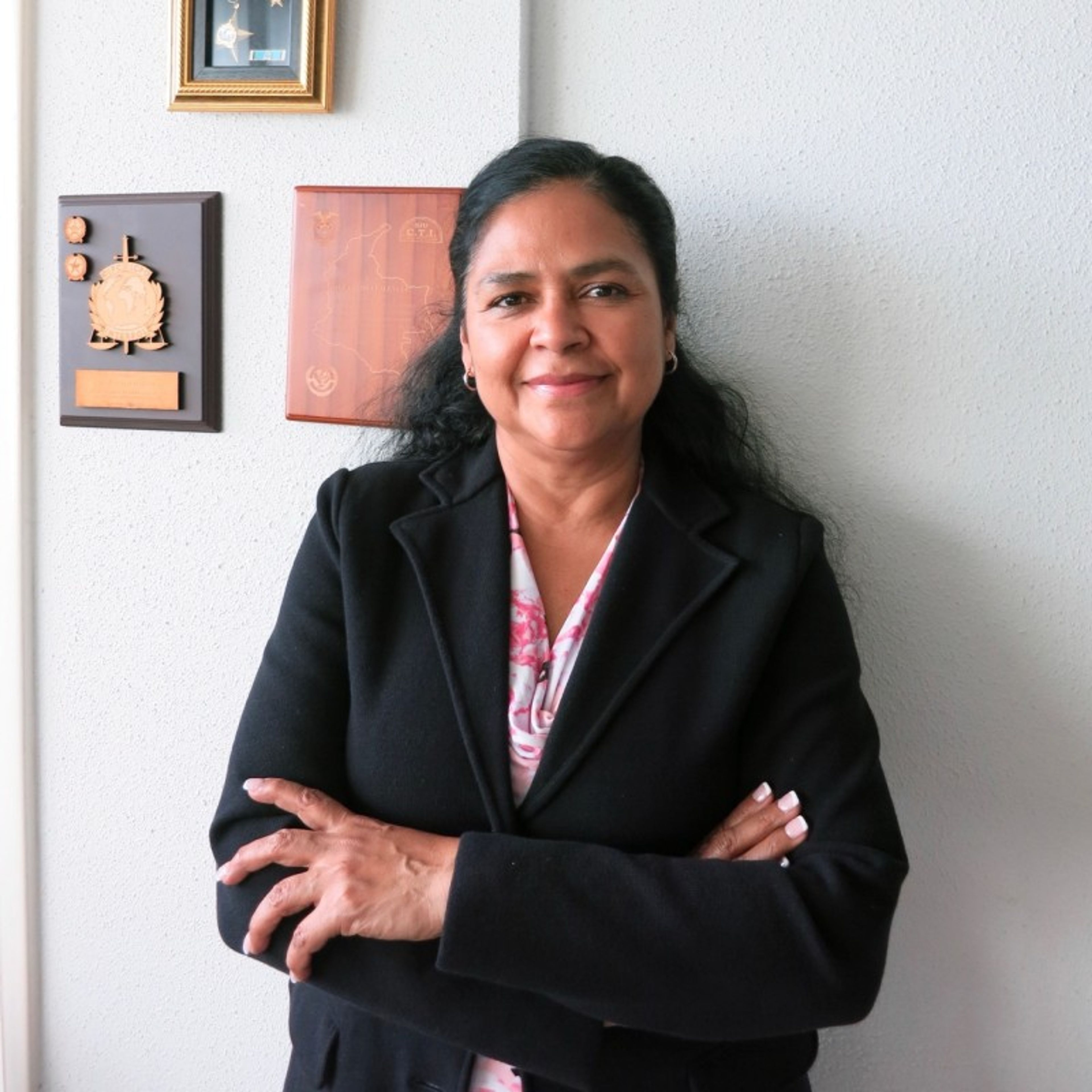 A woman with dark hair, wearing a black blazer and patterned blouse, smiles faintly with her arms crossed.