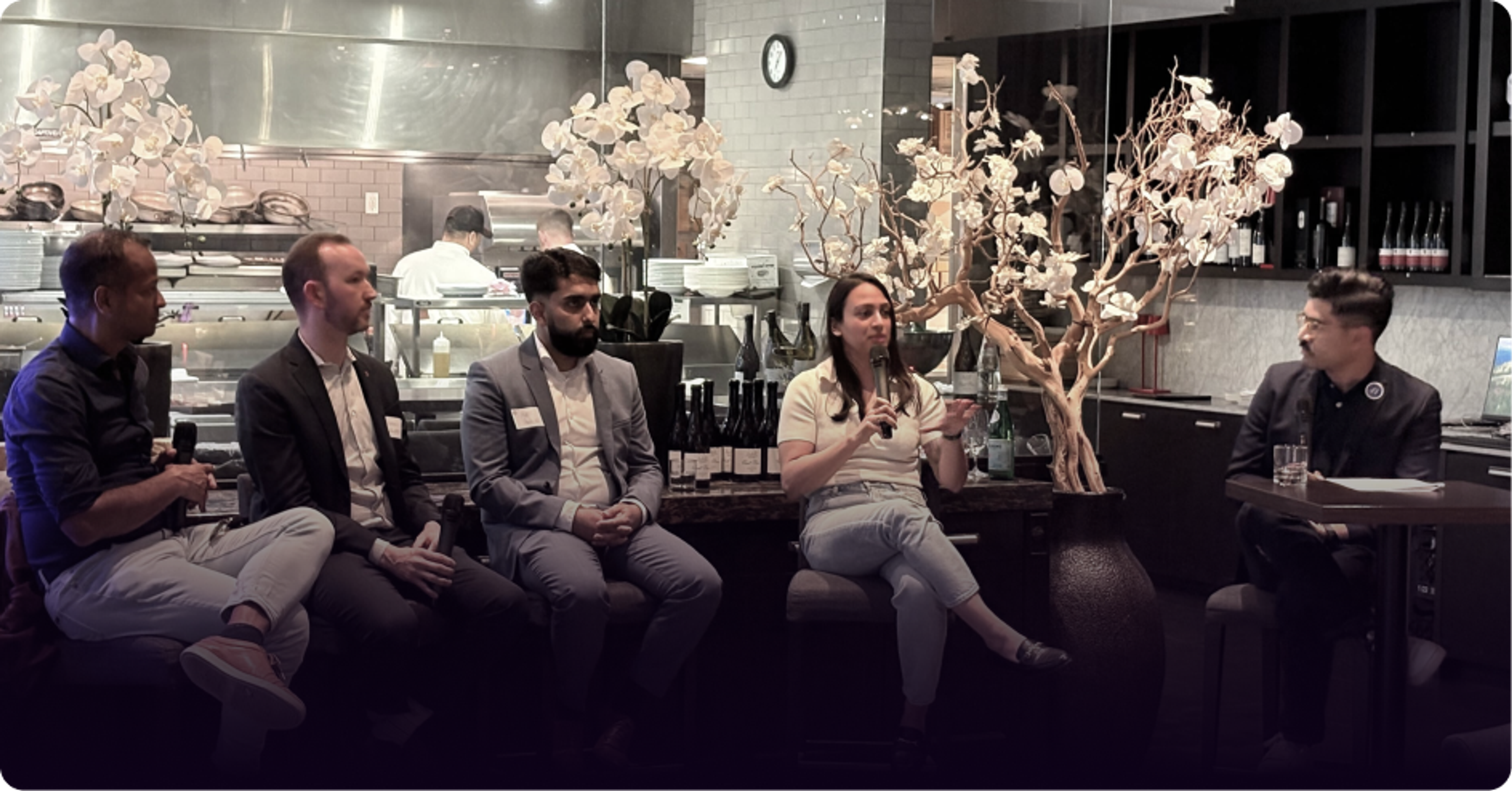A diverse panel of five people (four men, one woman) seated at a restaurant, with the woman speaking into a microphone.