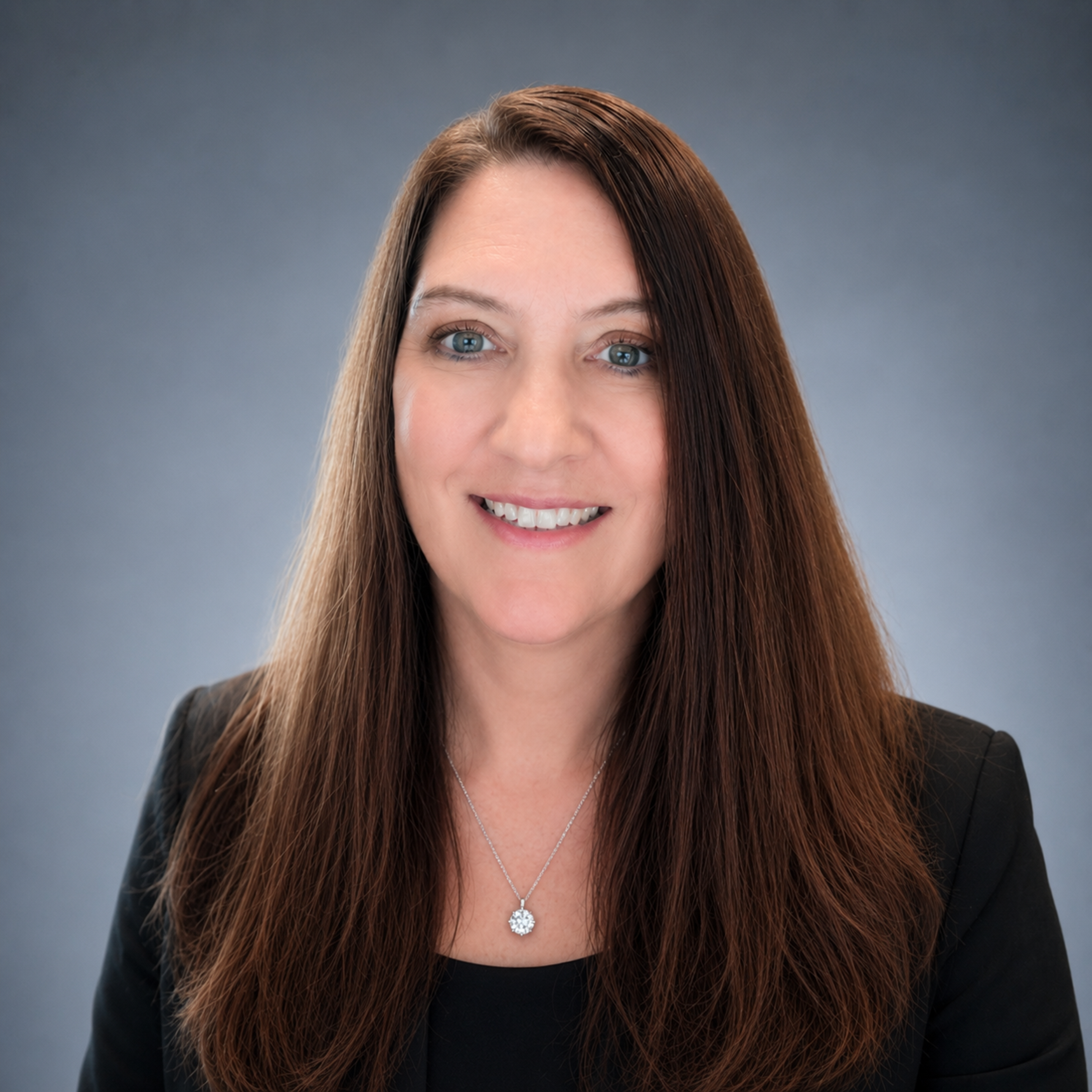 Smiling woman with long brown hair, blue eyes, wearing a black blazer and a silver necklace, against a grey background.