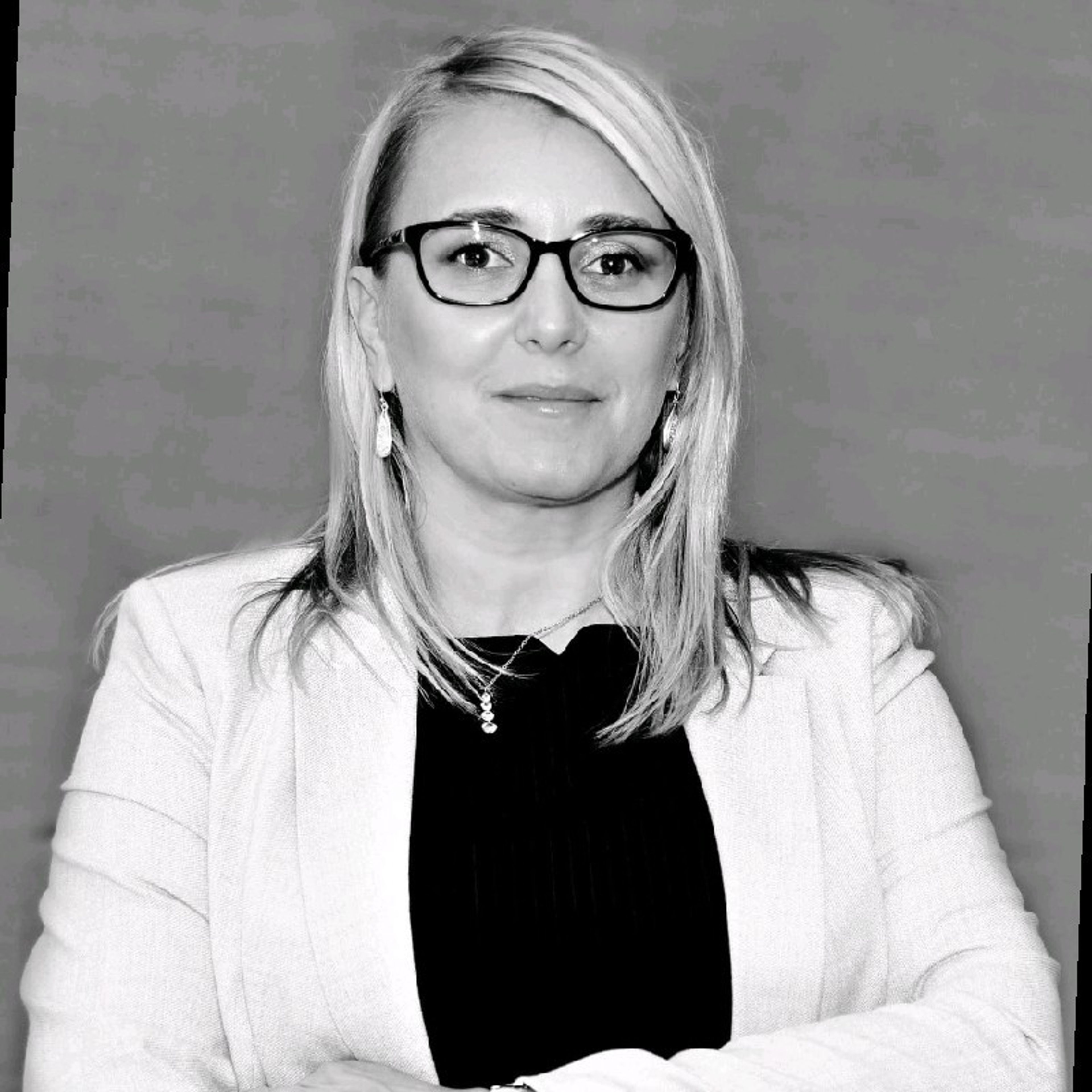 Black and white portrait of a woman with blonde hair, glasses, and a blazer, arms crossed.