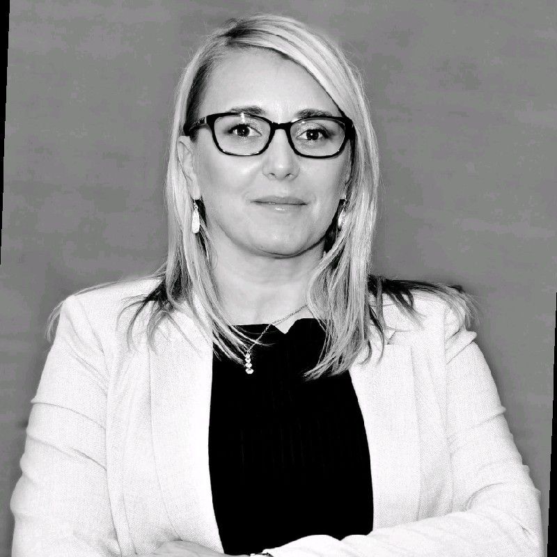 Black and white portrait of a woman with blonde hair, glasses, and a blazer, arms crossed.