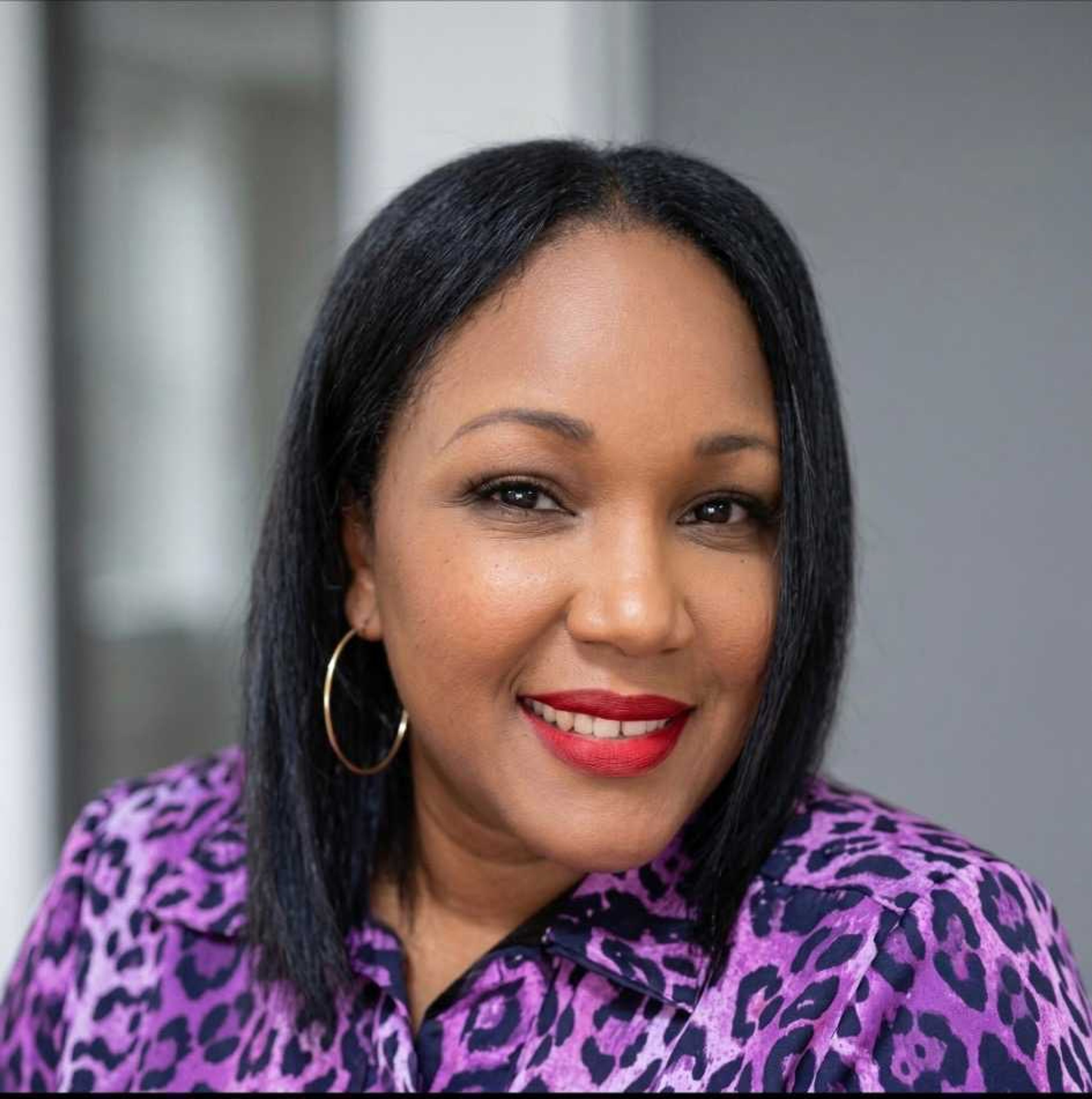 Headshot of a smiling woman with dark skin, dark hair, red lipstick, and a purple leopard print shirt.