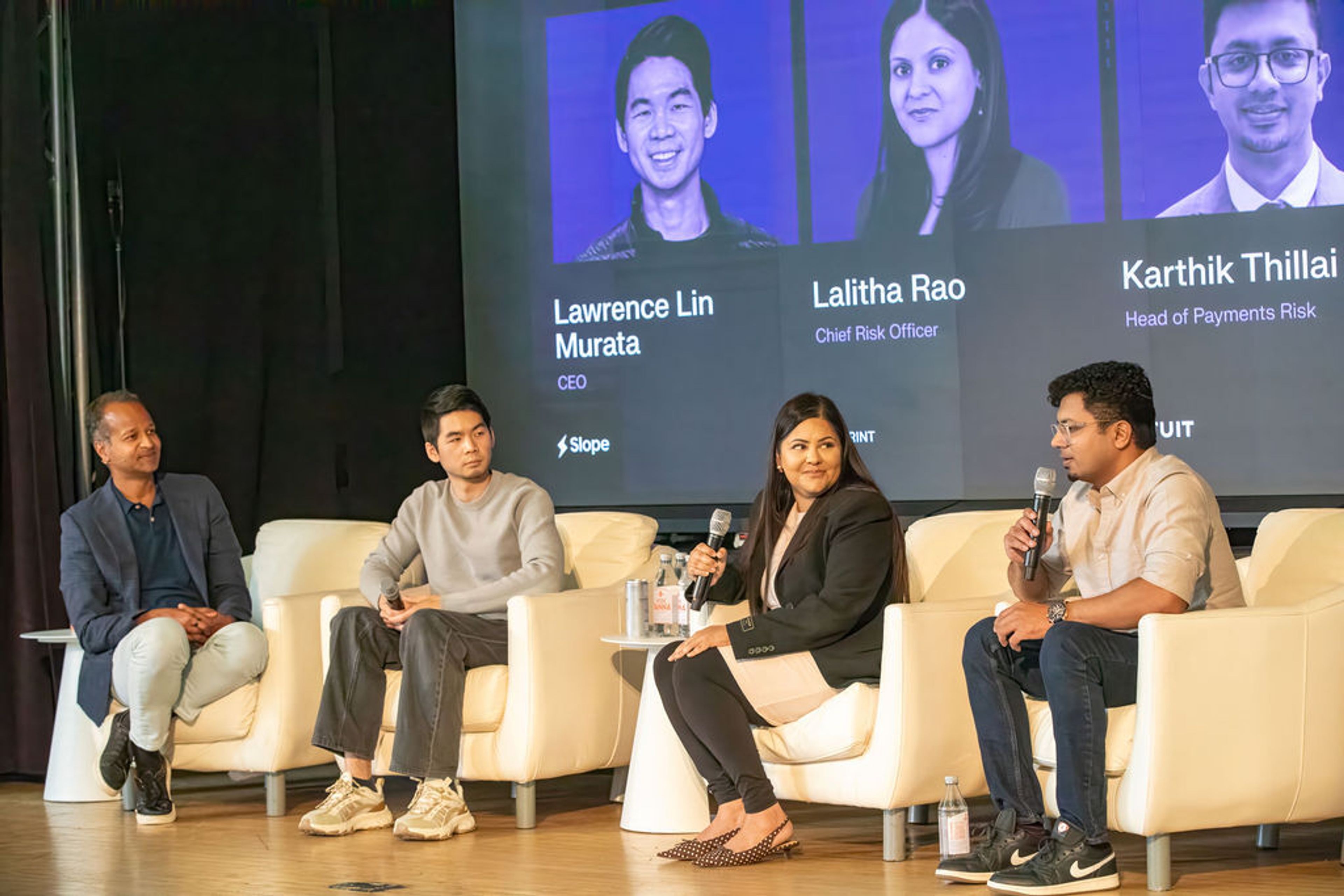 Four panelists sit on a stage, two holding microphones, with a screen displaying names and titles behind them.