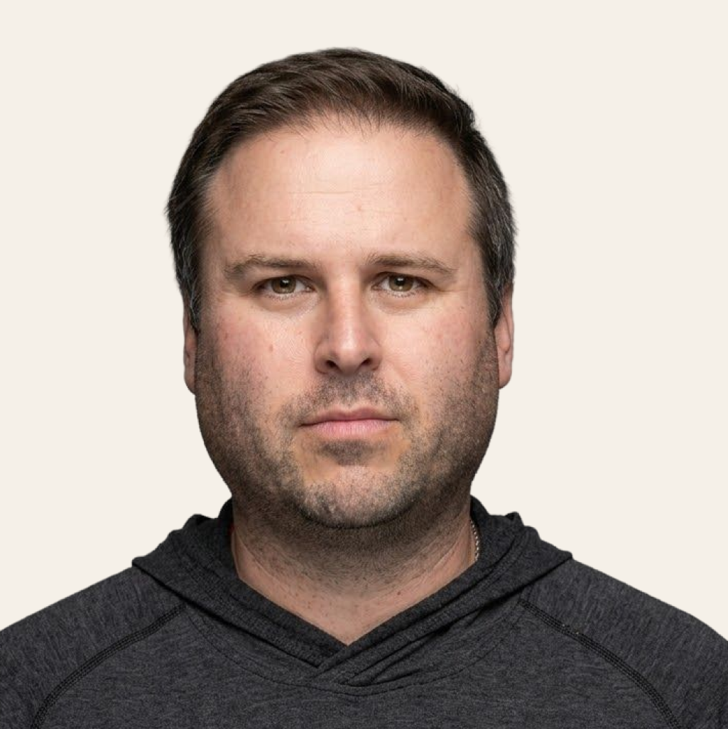 A man with short brown hair and light stubble wears a dark gray hoodie and looks directly at the camera.