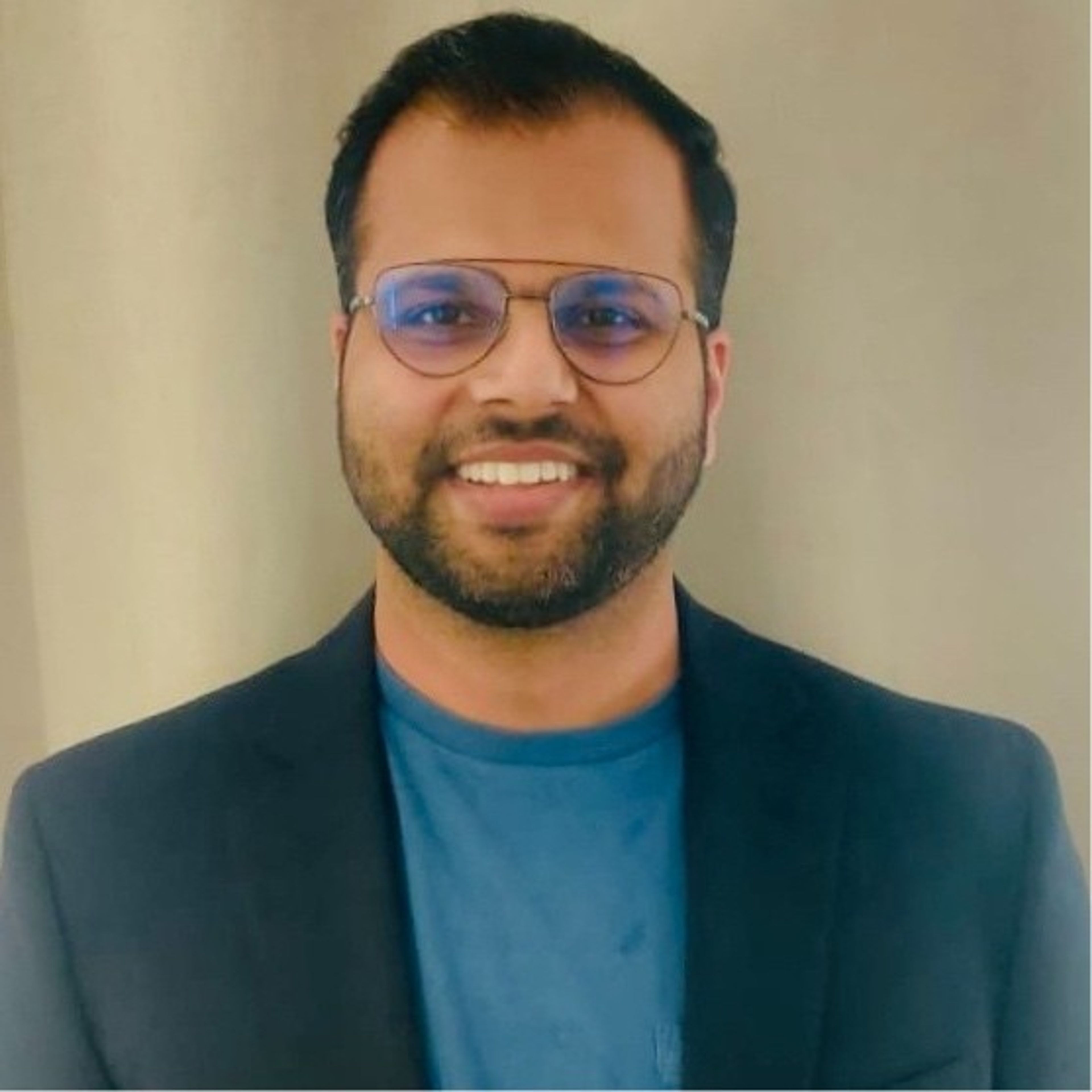 A smiling man with glasses and a beard, wearing a black blazer over a blue t-shirt.