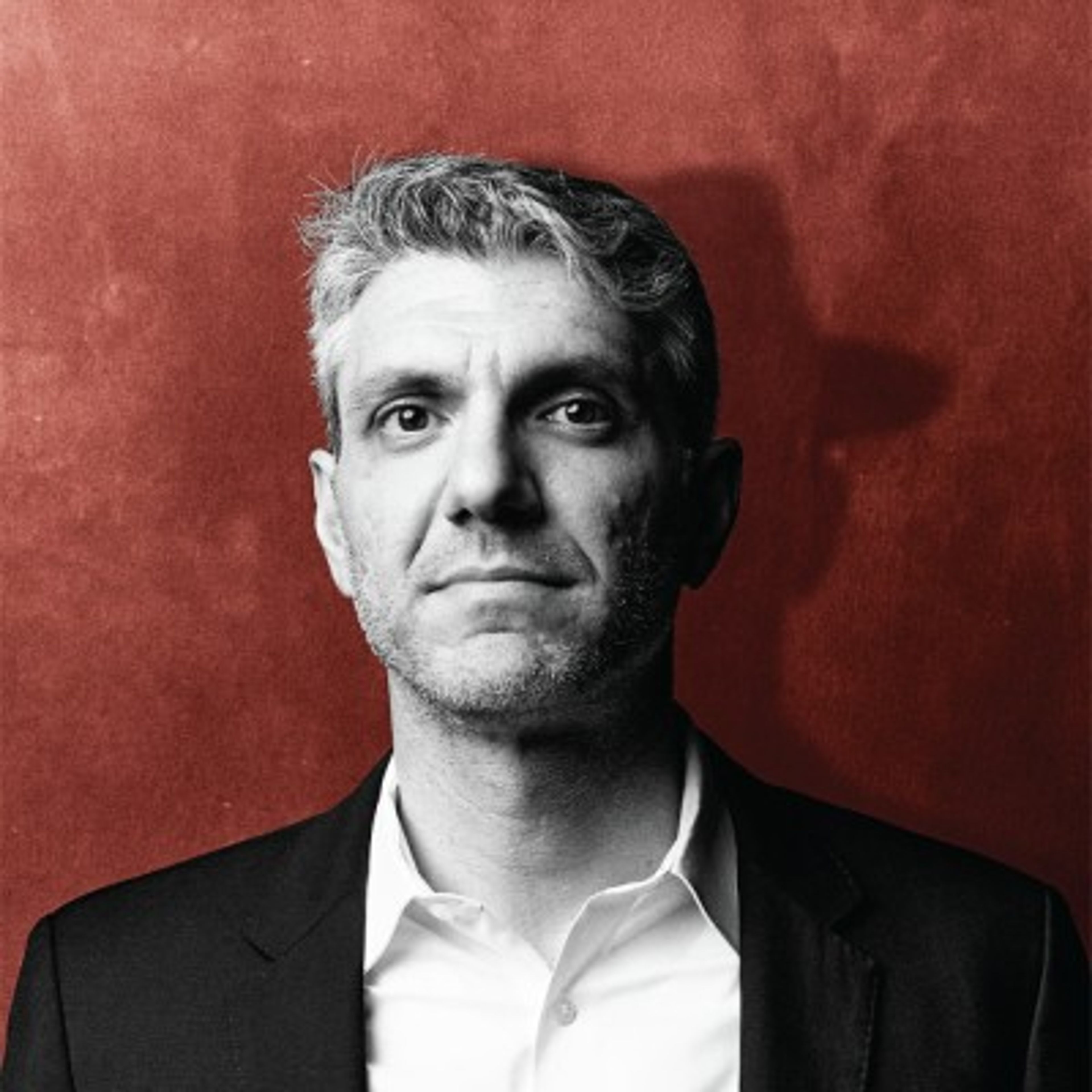 Portrait of a man with grey hair and light stubble, wearing a suit jacket and white shirt, against a reddish textured wall.