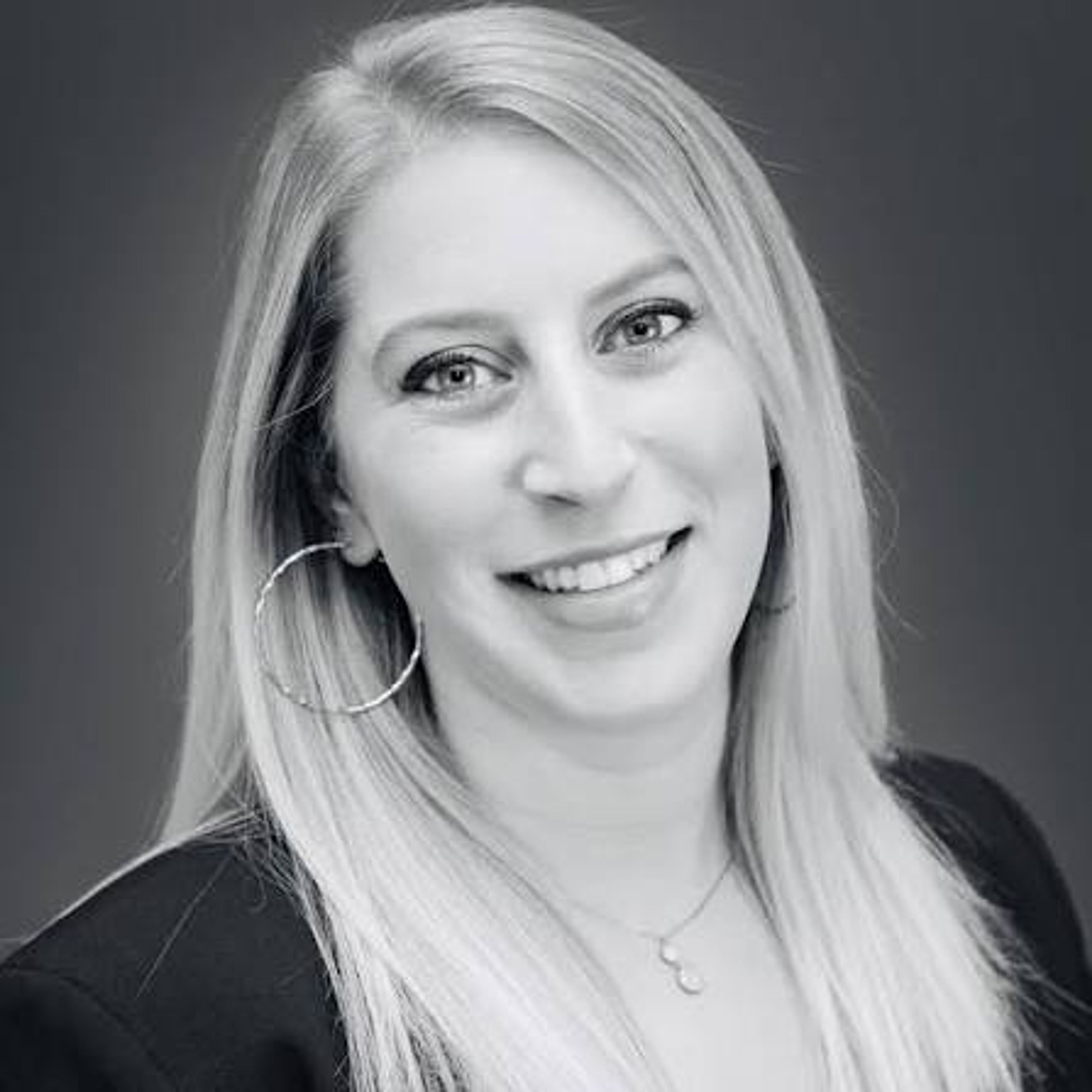 Black and white headshot of a smiling woman with long blonde hair, wearing hoop earrings and a necklace.