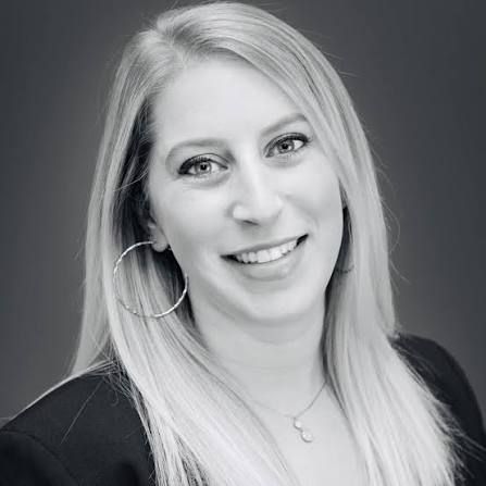 Black and white headshot of a smiling woman with long blonde hair, wearing hoop earrings and a necklace.