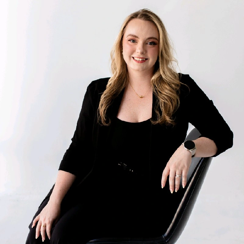 Smiling woman with long blonde hair wearing a black suit, sitting in a chair.