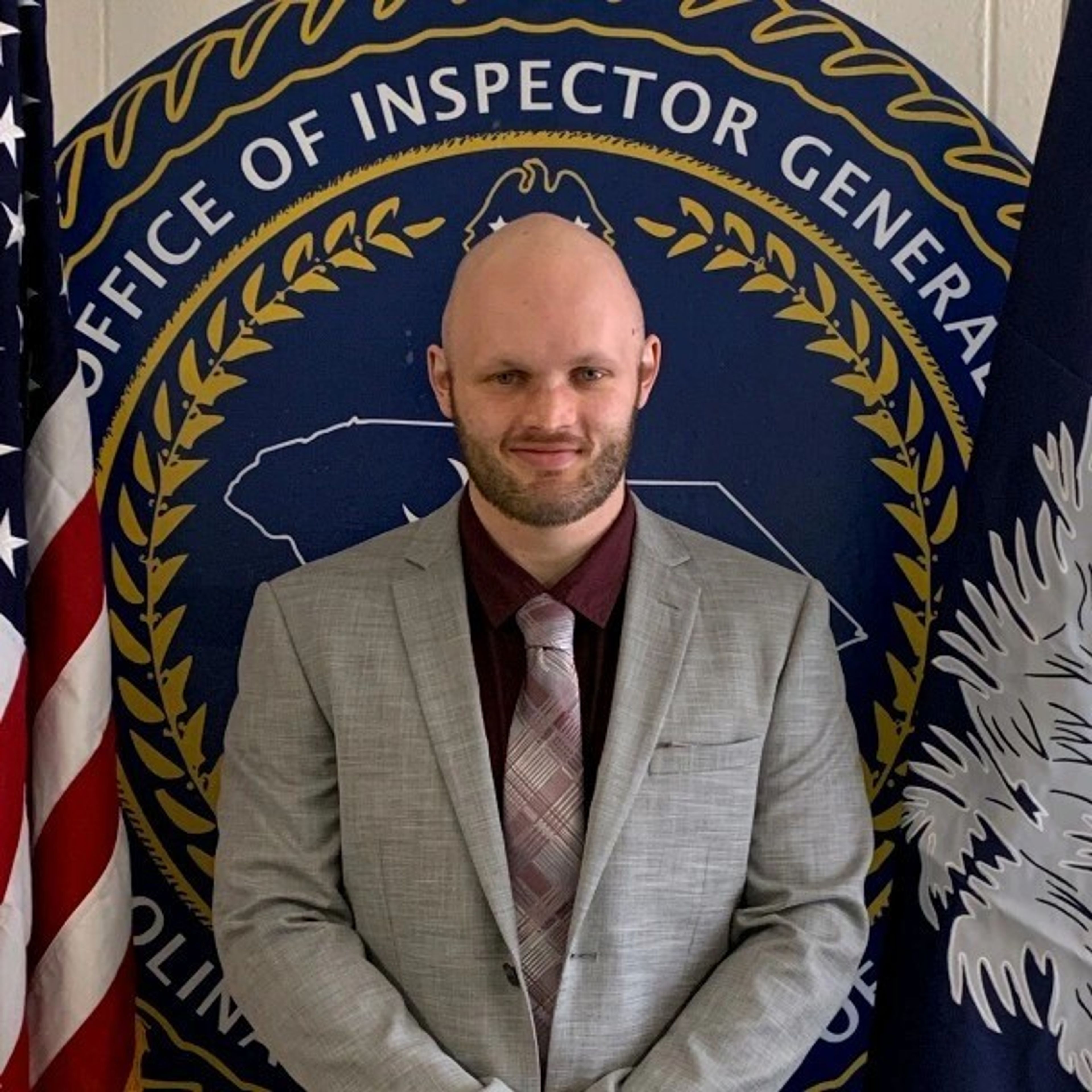 A smiling, bald man with a beard, wearing a gray suit and patterned tie, stands before the South Carolina Office of Inspector General seal, flanked by the US and South Carolina flags.