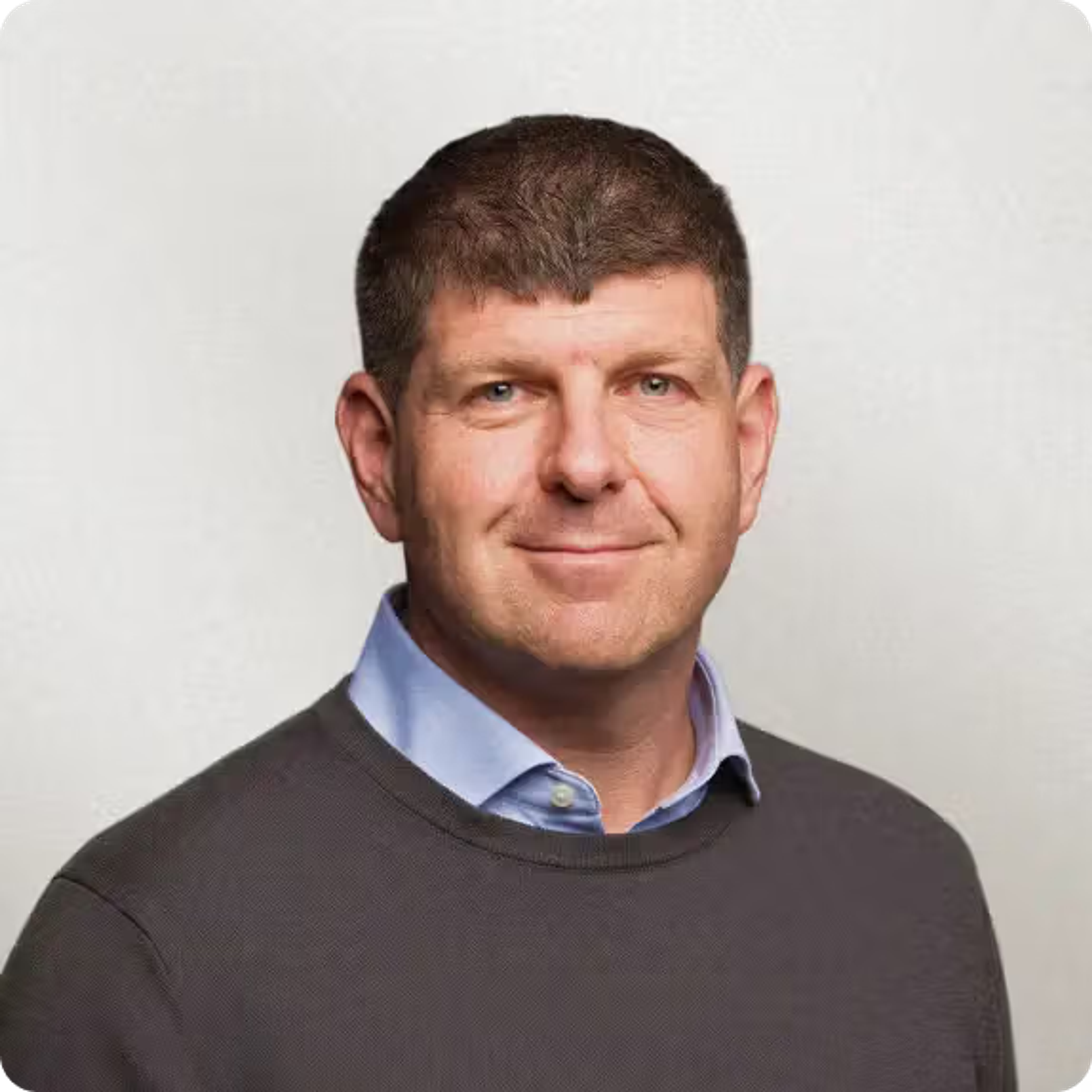 Headshot of a smiling middle-aged man with short brown hair, wearing a gray sweater over a blue shirt.