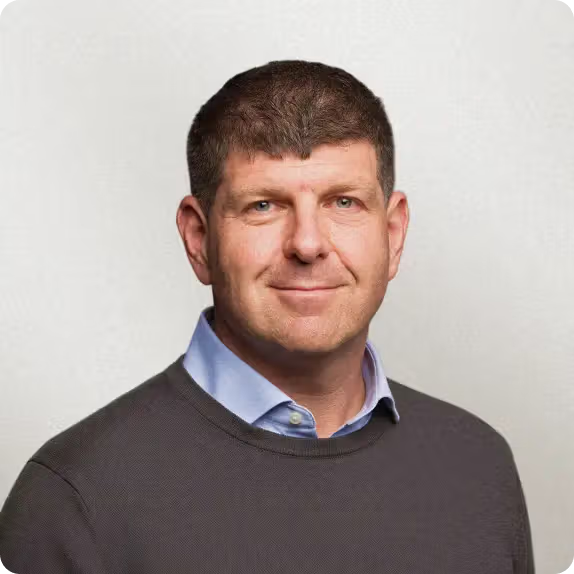 Headshot of a smiling middle-aged man with short brown hair, wearing a gray sweater over a blue shirt.