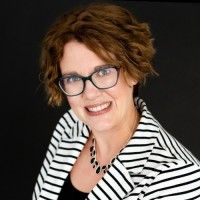 A smiling woman with short brown hair and glasses, wearing a black and white striped blazer.