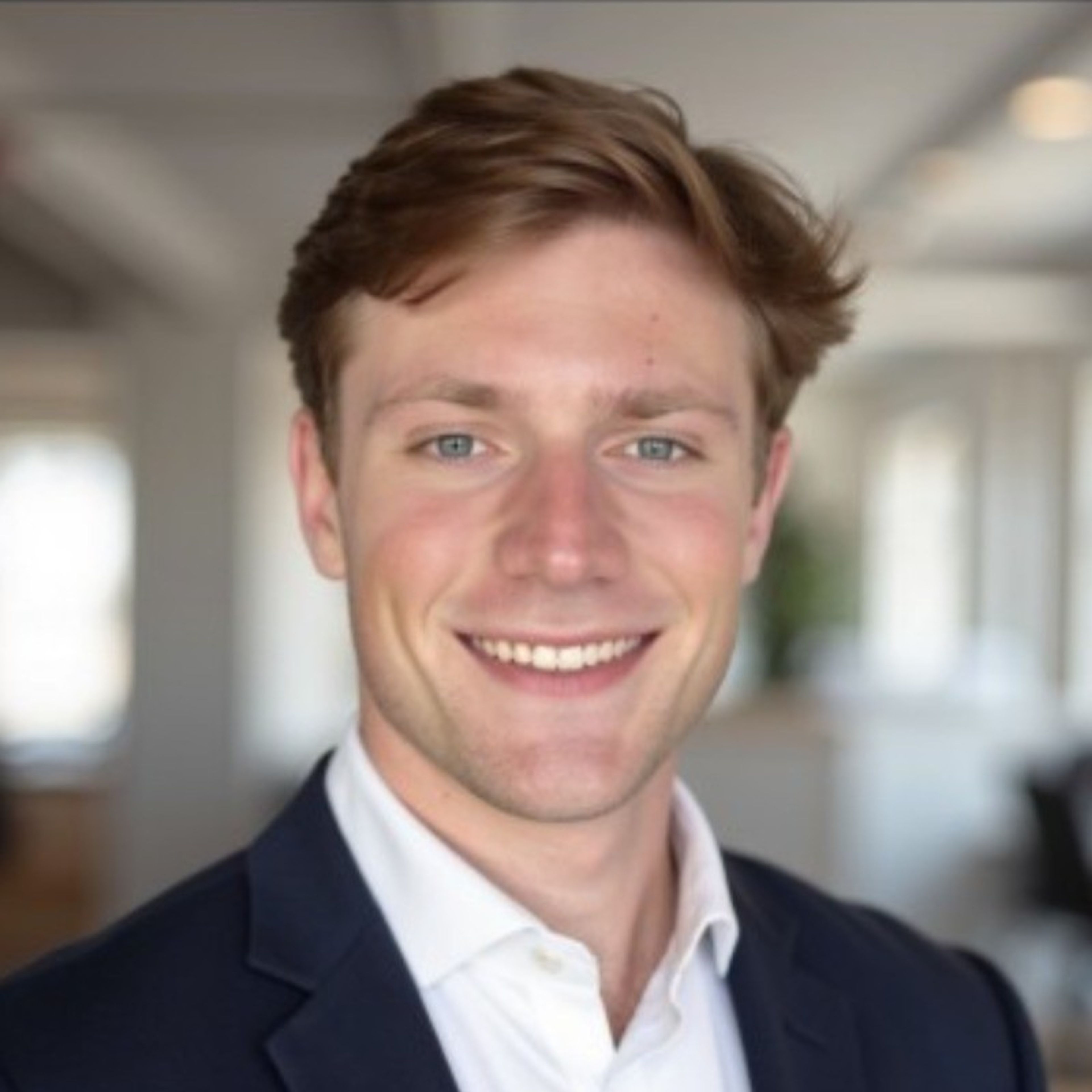 A smiling young man with light brown hair and blue eyes, wearing a navy suit jacket and white shirt.