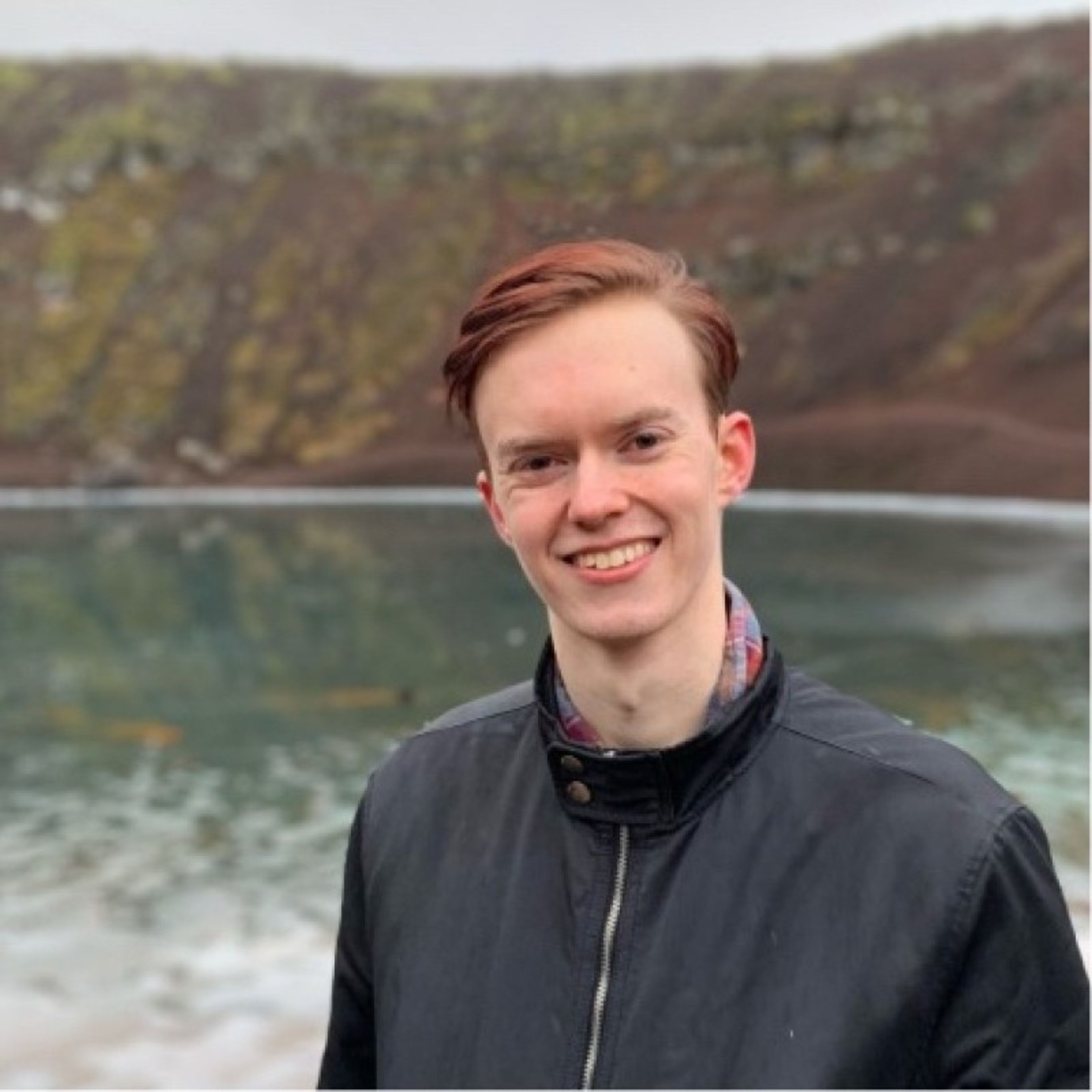 A smiling young man with reddish hair wearing a black jacket stands in front of a crater lake in Kerið Crater, Iceland.