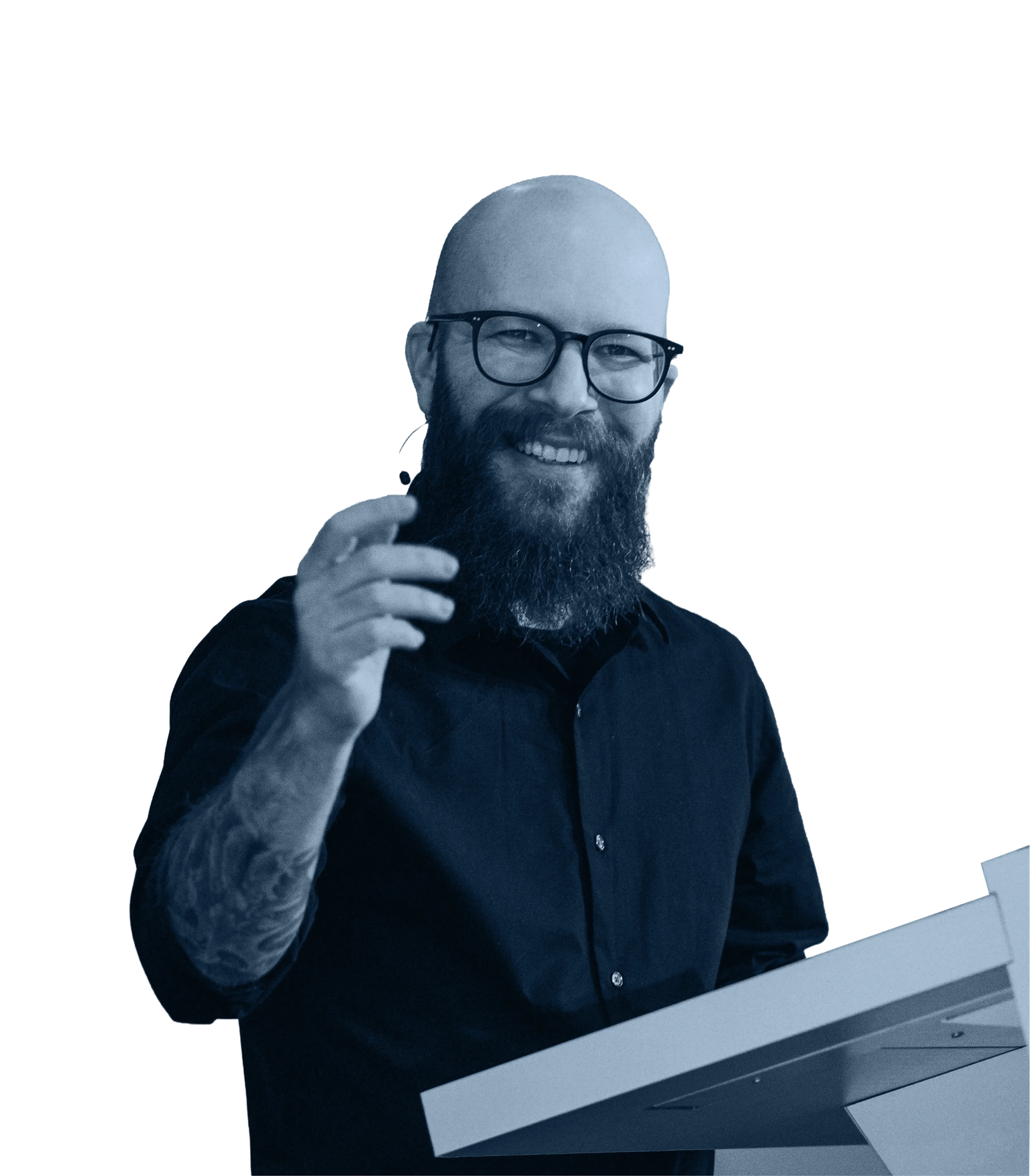 Smiling bald man with a long beard, glasses, and earpiece microphone stands at a podium.