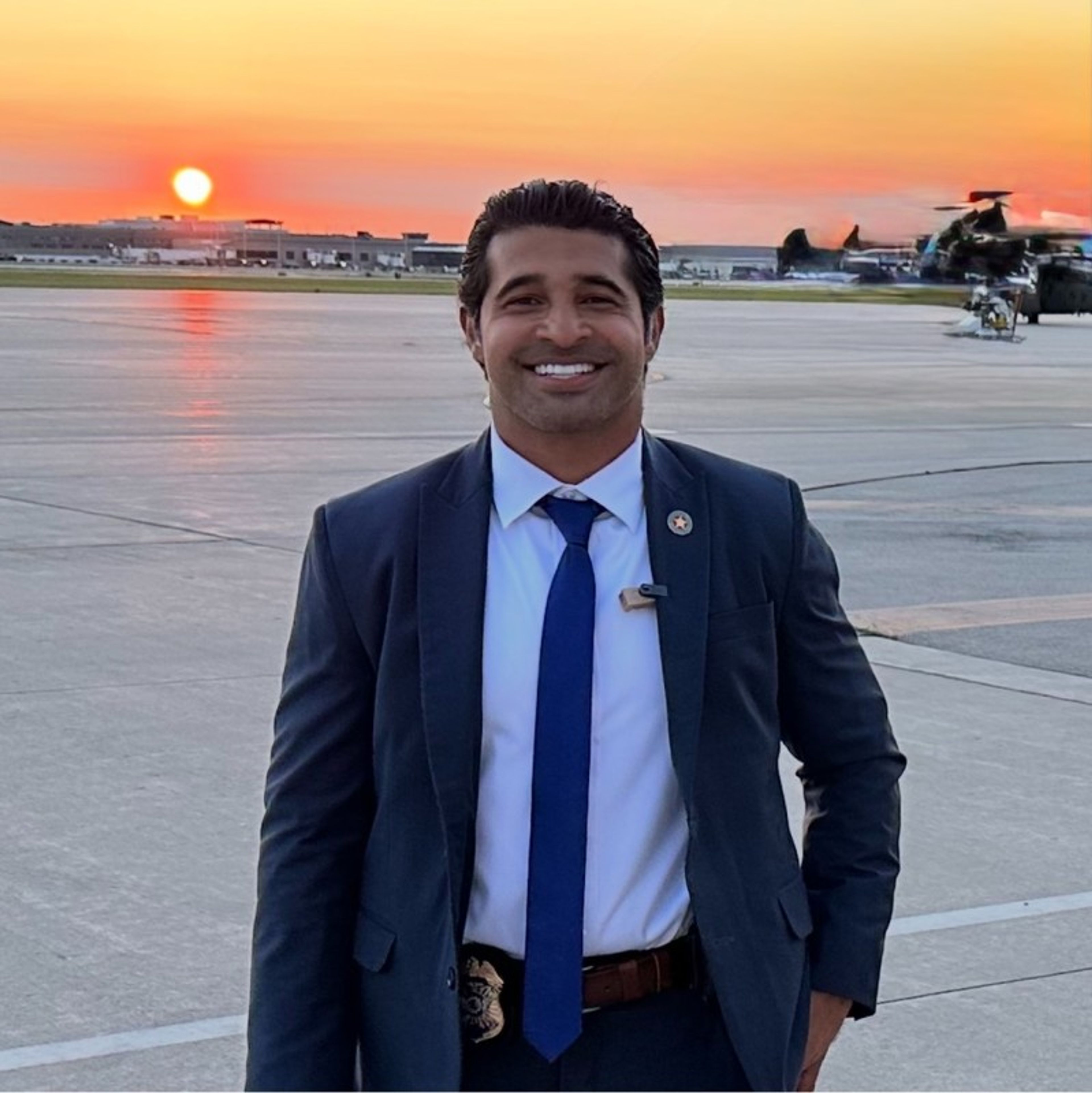 Smiling man in a suit with a badge on an airport tarmac at sunset.